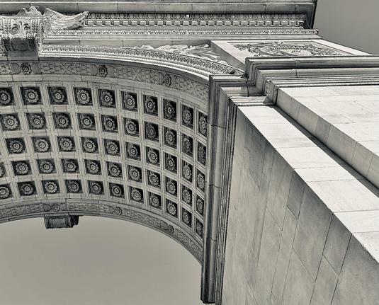A photograph of the Washington Square Arch in New York City. The perspective is from beneath and slightly to the side of the arch's main barrel vault, looking steeply upward. The image captures the intricate coffered ceiling of the archway, which is decorated with a repeating pattern of small rosettes within square panels. On the right side of the image, the massive stone pier that supports the arch rises, showcasing its smooth, large blocks of masonry and a pronounced cornice detail at the upper-right edge. The sky visible through the arch is a bright, featureless expanse, a result of the high-contrast processing I did for artist effect, and which makes the white marble of the structure stand out strongly.