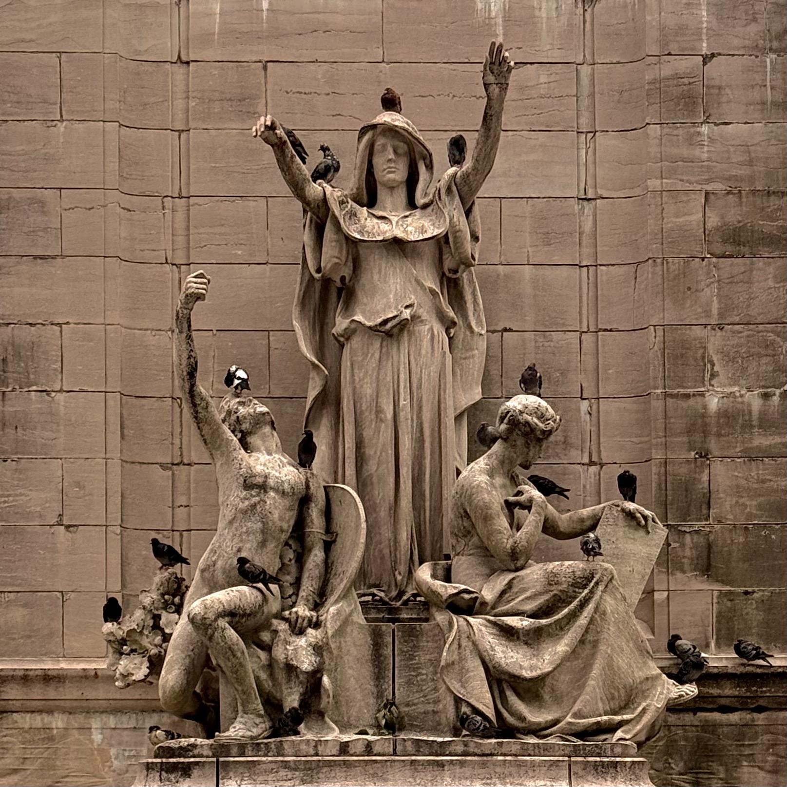 A color, sepia-toned photograph taken on an overcast afternoon of a stone sculpture on the U.S.S. Maine National Monument featuring three allegorical figures: a central robed woman with raised arms, a seated man with an uplifted arm, and a seated woman looking at a scroll. Many pigeons are perched on the figures and monument.