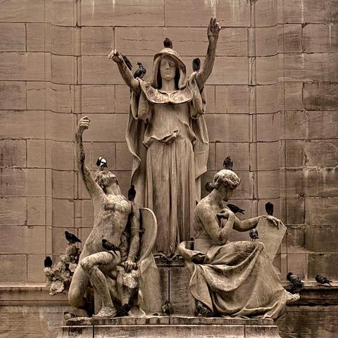 A color, sepia-toned photograph taken on an overcast afternoon of a stone sculpture on the U.S.S. Maine National Monument featuring three allegorical figures: a central robed woman with raised arms, a seated man with an uplifted arm, and a seated woman looking at a scroll. Many pigeons are perched on the figures and monument.