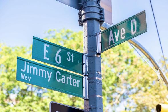 A color photograph of a close-up, outdoor shot of a metal utility pole at an intersection holding street signs against a bright blue sky with green and yellow foliage from trees in the background. The signs are stacked and mounted to the pole with metal brackets. They are green with white lettering. The visible street signs read: "Ave D" (top right), "E 6 St" (middle left), and "Jimmy Carter Way" (bottom left). A small, black and white one way traffic sign is partially visible at the very bottom center. It is mid morning on a clear day. Photo courtesy of EV Grieve.