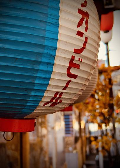 A color photograph in portrait orientation. A close-up, slightly low-angle photograph of a large, cylindrical Japanese paper lantern. The lantern is primarily white and blue. The left half is solid teal-blue, and the right half is white with large, bold, red Japanese characters written vertically down the center in Katakana script. The lantern has a bright red base and. A small black metal ring is visible attached to the red base. The paper texture of the lantern is ribbed. The background is softly blurred, featuring warm, golden-yellow bokeh lights and indistinct shapes of the market in the background. Another similar lantern, slightly out of focus, is visible behind the main one.