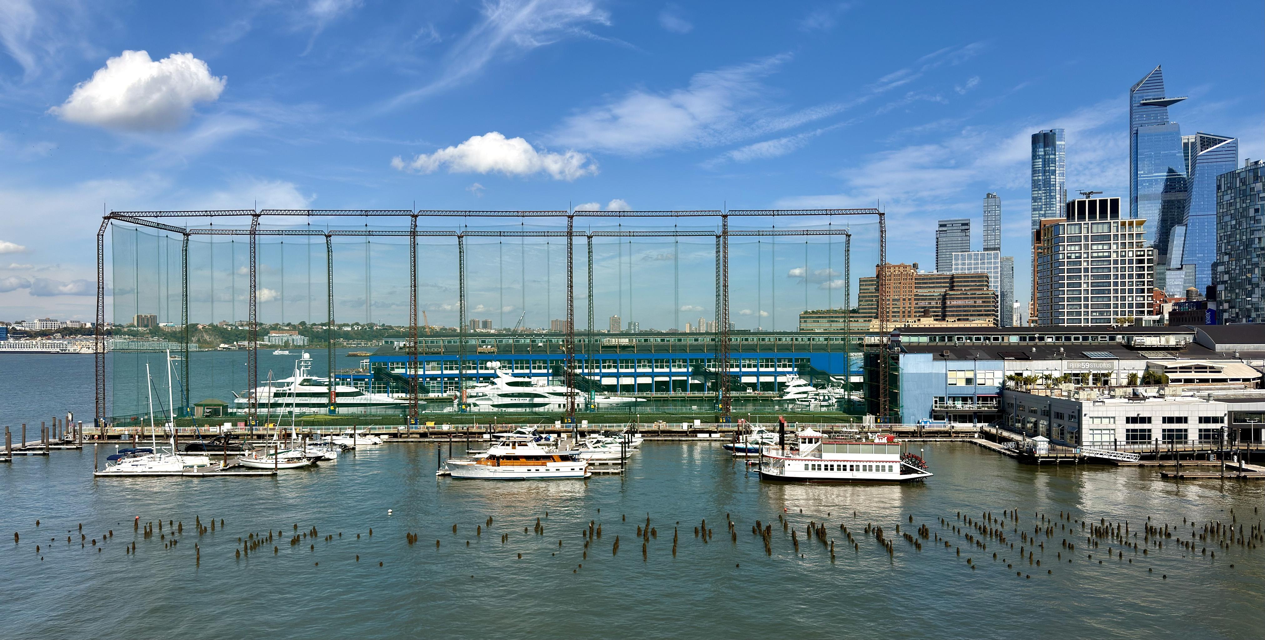 A color, wide-angle photograph taken midday captures a view across the water from Pier 57 toward Chelsea Piers in New York City. The focal point is the massive, caged structure of the golf club driving range, which is a tall, glass-walled building enclosed in a fine-mesh netting structure. The building's glass façade reflects the water, sky, and surrounding cityscape. In the foreground, the water of the Hudson River is choppy and a bit murky, with numerous old, wooden pier pilings sticking up from the water. A small marina area is visible with numerous white boats docked, including a few yachts and a ferry-style boat. To the right of the golf structure, a large, dark blue/gray building, part of the Chelsea Piers complex, is visible at the edge of the water. In the background, beyond the pier complex, modern skyscrapers of the Hudson Yards neighborhood dominate the skyline, including the distinctive, angular tops of several tall glass towers. The sky is bright blue with a few puffy white clouds.