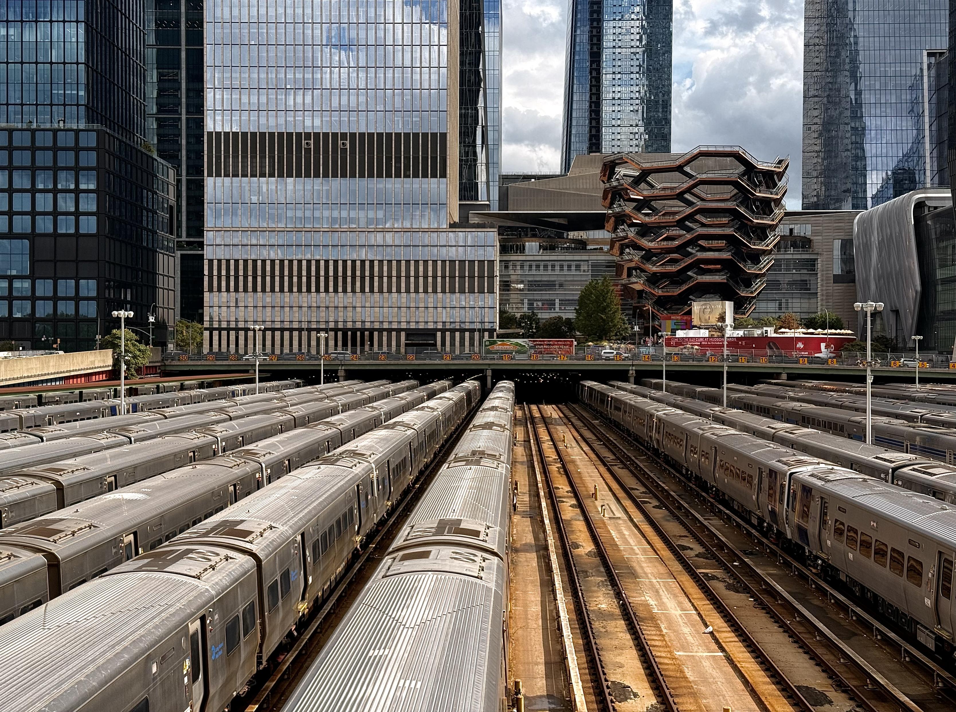 A color photograph of the the expansive West Side Yard rail depot in Manhattan on a partly cloudy afternoon, with the modern architectural structures of Hudson Yards rising directly behind it. In the foreground and midground, dozens of silver passenger trains are parked on parallel tracks, filling the width of the frame. The trains are lined up tightly in sections of the yard. The tracks run horizontally into the distance, with a central gap of exposed tracks and gravel creating a clear middle lane between the parked trains. Dominating the background are the tall, glass and steel skyscrapers of the Hudson Yards development. Prominently featured, just above the trains, is the unique, honeycomb-like, copper-colored structure known as The Vessel. Other sleek, dark, and reflective office and residential towers surround it.