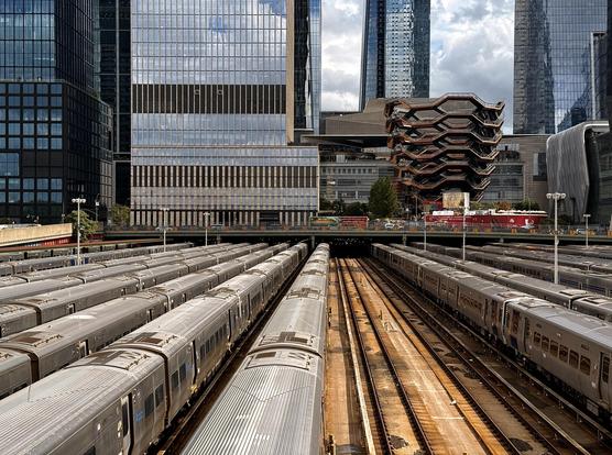 A color photograph of the the expansive West Side Yard rail depot in Manhattan on a partly cloudy afternoon, with the modern architectural structures of Hudson Yards rising directly behind it. In the foreground and midground, dozens of silver passenger trains are parked on parallel tracks, filling the width of the frame. The trains are lined up tightly in sections of the yard. The tracks run horizontally into the distance, with a central gap of exposed tracks and gravel creating a clear middle lane between the parked trains. Dominating the background are the tall, glass and steel skyscrapers of the Hudson Yards development. Prominently featured, just above the trains, is the unique, honeycomb-like, copper-colored structure known as The Vessel. Other sleek, dark, and reflective office and residential towers surround it.