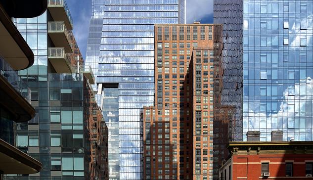 A color photograph in landscape orientation taken along the High Line in Manhattan on a partly cloudy day. A detailed, urban architectural collage showing a mix of modern glass skyscrapers, mid-rise brick residential buildings, and a smaller, older brick structure at the bottom right, all tightly packed together. The scene is dominated by contrasting vertical lines, textures, and reflections.