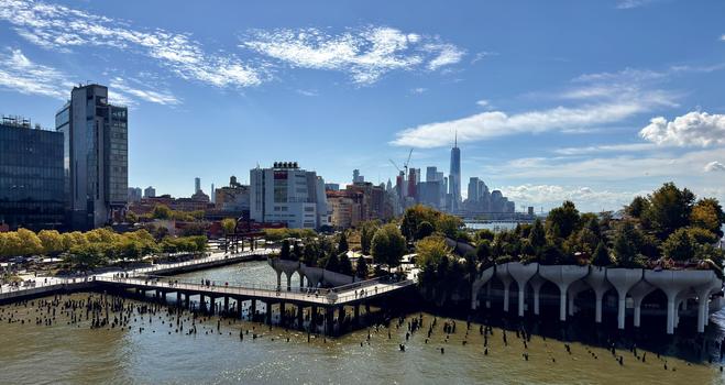 A color, wide-angle landscape photograph, an elevated shot showing the pier scene along the Hudson River in Manhattan on a sunny afternoon. The foreground is dominated by Little Island at Pier 55, an artificial public park supported by a unique structure of white, mushroom-like concrete columns. The park's surface is lush with green trees and vegetation. A wooden walkway bridge connects the island to the mainland (left side of the frame). The water surrounding the pier and the island is a murky, dark color, with numerous decaying, dark wooden piles sticking out of the water. In the midground, beyond Little Island and the waterfront, the Meatpacking District/Chelsea area is visible with modern glass buildings on the far left and older, red and white brick industrial buildings and rooftops. The background features the Lower Manhattan skyline, dominated by the distinctive spire of One World Trade Center standing tall among other high-rise buildings. People are visible walking on the bridge and on the island. The sky is a bright blue with scattered white, fluffy clouds. 