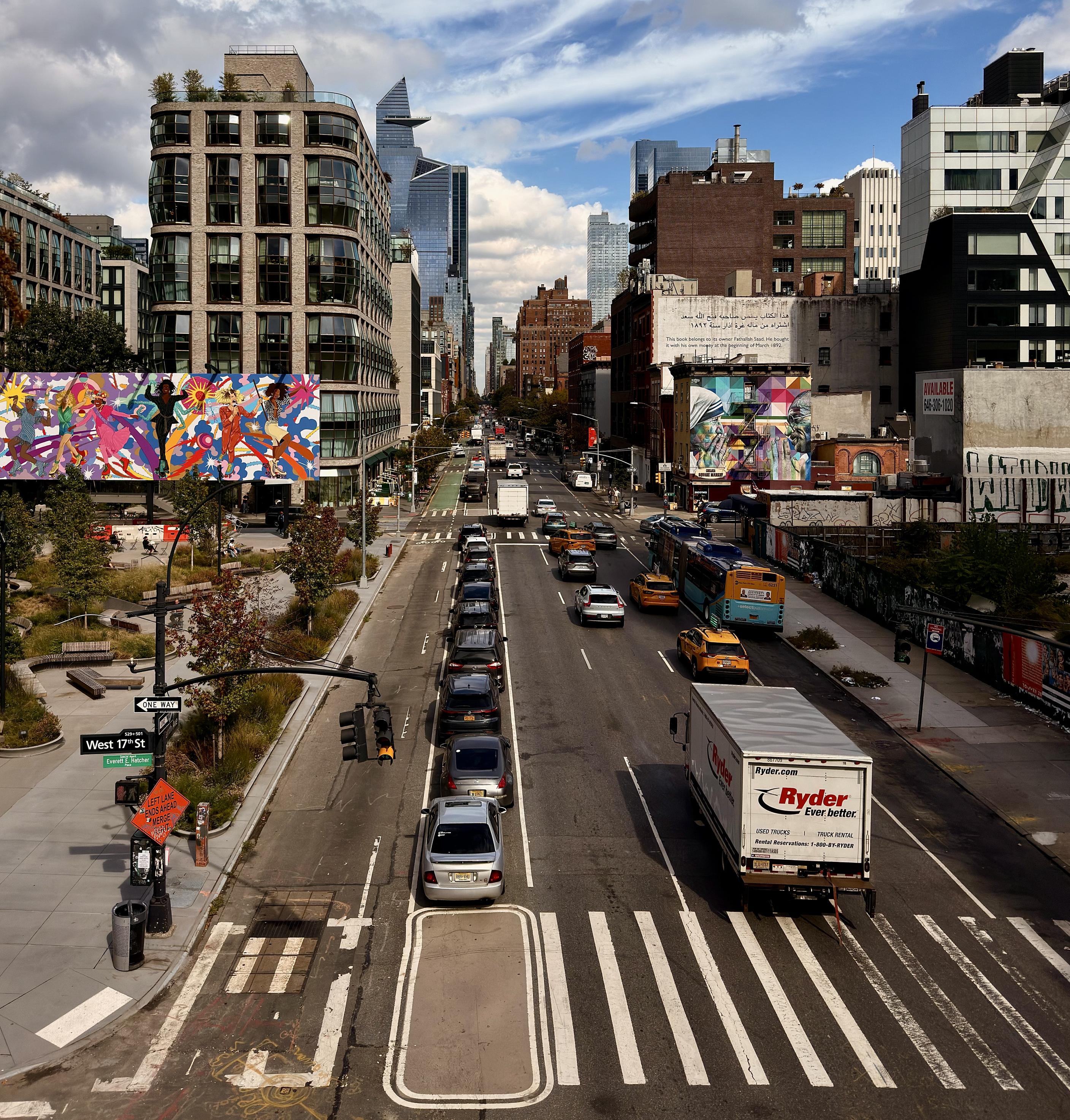 A color photograph in square aspect ratio. A street view of the busy Tenth Avenue in the Chelsea neighborhood of Manhattan taken from the High Line elevated park on a mostly clear afternoon. The street runs toward the background, lined on both sides by diverse urban architecture. In the foreground on the left, there is a section of the High Line park, featuring trees and wooden benches. Directly behind this park section is a prominent modern residential building with a dark brick facade. A large-scale colorful canvas artwork is mounted in front of this building. Behind the art, the building's distinctive curved or 'bubble' bay windows are visible, giving the structure a unique, textured appearance. Further down the street, another building with an unusually tapered curved glass facade is visible on the far left, standing out against the sky. The right side of the street is lined with commercial buildings and billboards, featuring bright advertisements. A large white truck from Ryder is prominently positioned in the right foreground. The multi-lane avenue is busy with traffic, including several parked cars, a long line of vehicles stopped at a light, yellow taxis, and a blue and yellow public bus. A crosswalk and a street sign for West 17th Street are visible in the lower left corner. The background features a classic New York city skyline with a mix of older brick buildings and modern glass skyscrapers.