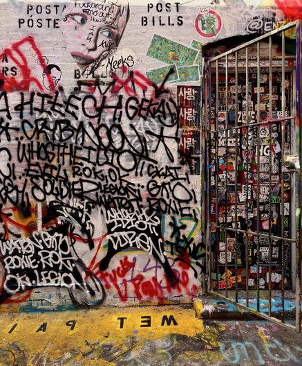 A color photograph of a brick wall in an alley densely covered in layers of street art and vandalism on a sunny day. The wall itself is light gray. The surface is completely covered in black and red graffiti tags, with large, looping black script dominating the center and left, making individual words difficult to distinguish. Near the top-left, a mural portrait of a stylized person’s face is partially visible and tagged over. Red spray paint streaks are prominent on the left side of the wall. To the right, a weathered metal security gate stands open, slightly ajar. The gate and the wall panel it opens onto are absolutely covered in a dense collage of small stickers, featuring various designs, logos, and smaller pieces of street art. The ground in the foreground is rough pavement with patches of yellow paint, and the word "WET PAINT" is painted in large, inverted black letters near the bottom center.