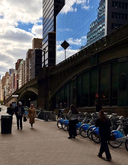 A color photograph in portrait orientation. The composition features a Citibike station lined up on the right side of a wide, paved sidewalk. A few pedestrians are walking along the sidewalk: a man and a woman in the foreground on the left, and several people near the bike rack on the right. Dominating the center of the image is a dark, arched, elevated structure, for an overpass, that runs diagonally from the center-right to the middle-left. Above and behind this structure, the glass facade of a tall, modern skyscraper reflects the partially cloudy blue sky and the structure itself. To the left, a row of older, mixed-height buildings with varying brick and stone facades recedes into the distance. The lighting shows it's a bright day with scattered clouds, casting a dramatic play of light and shadow, with the lower sidewalk area in partial shadow from the overhead structure.