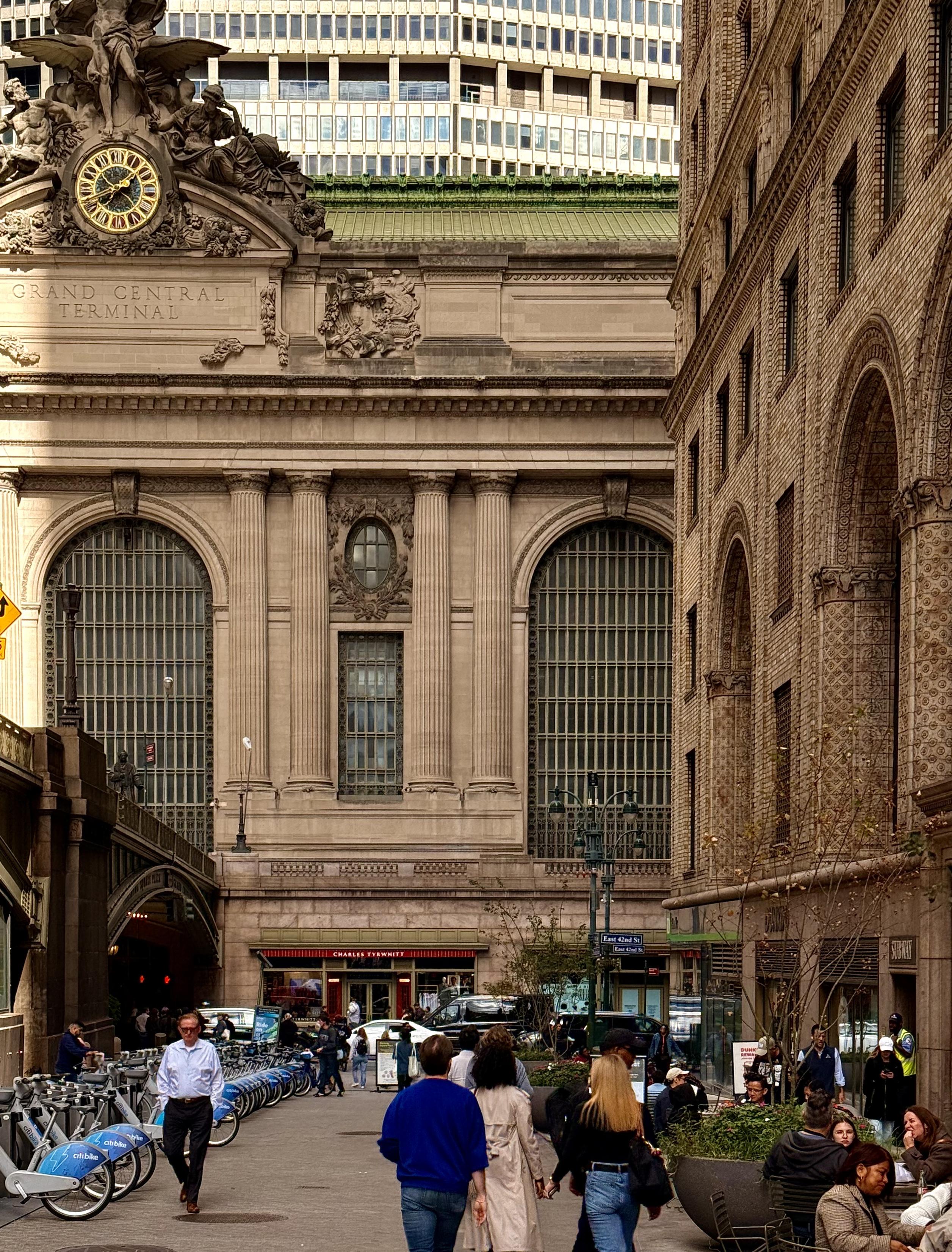 A color photograph of the large, ornate, light-tan Beaux-Arts facade of the Grand Central Terminal building dominates the center and background. The top of the building features a large clock and classical sculptural elements, including the famous 13-foot-wide Tiffany clock. Below the sculptural elements, the words "GRAND CENTRAL TERMINAL" are carved into the stone. The building has three prominent, large arched windows with visible internal grid patterns, separated by Corinthian columns. To the right, a multi-story building of darker, reddish-brown brick stands at an angle, featuring multiple arched windows and giving the impression of classic New York architecture. In the foreground, a group of pedestrians is walking towards the viewer on a paved walkway. A man in a light blue shirt and dark pants is walking toward the left foreground. Closer to the right, a small group of people are walking away from the camera, including a man in a blue hoodie and a person in a light trench coat. To the far left, a row of shared rental bicycles is parked along the edge of the walkway. Further in the distance, a modern, light-colored skyscraper with many vertical lines rises above the Terminal, contrasting with the classical architecture below. The lower part of the Terminal's facade features a street level with some storefronts and traffic.