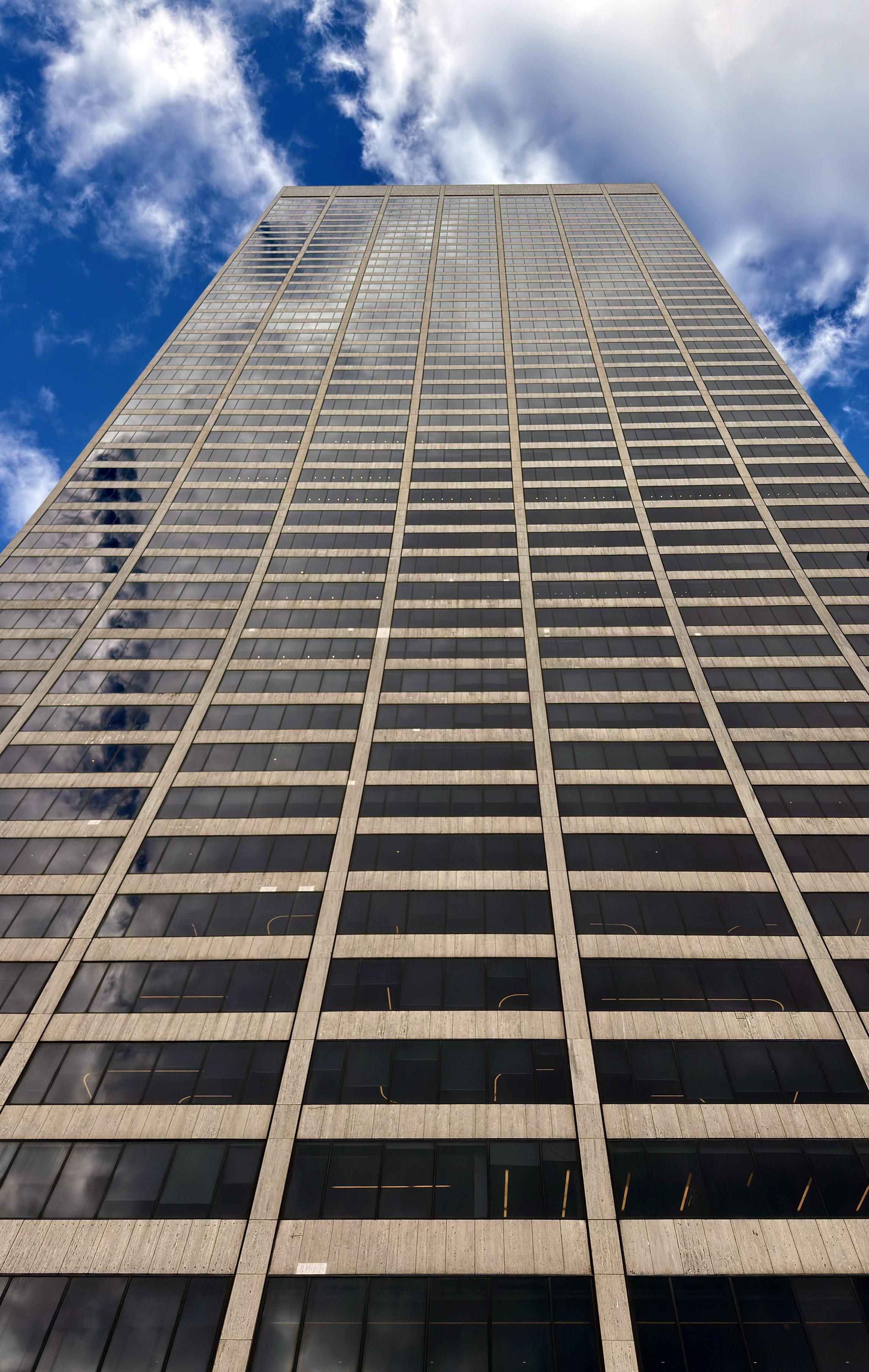 A color, low-angle, vertical shot the tall, modern skyscraper known as the Grace Tower. The tower is a grid of dark, reflective glass windows separated by light-colored, narrow vertical and horizontal concrete or stone mullions. The dark glass reflects the bright blue sky and white, wispy clouds above. The perspective is dramatically exaggerated, emphasizing the building's height as it tapers toward the top and seemingly disappears into the clouds. The lower part of the building fills the bottom of the frame, with the sky and clouds dominating the top portion.