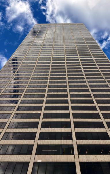 A color, low-angle, vertical shot the tall, modern skyscraper known as the Grace Tower. The tower is a grid of dark, reflective glass windows separated by light-colored, narrow vertical and horizontal concrete or stone mullions. The dark glass reflects the bright blue sky and white, wispy clouds above. The perspective is dramatically exaggerated, emphasizing the building's height as it tapers toward the top and seemingly disappears into the clouds. The lower part of the building fills the bottom of the frame, with the sky and clouds dominating the top portion.
