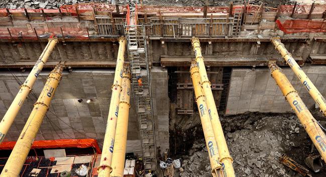 A color photograph in landscape orientation of an active construction site viewed from above on an overcast morning. The most prominent features are a series of massive, slanted yellow steel pipes running diagonally from the foreground toward the background structure. There are eight visible in total. In the background, there is a deep excavation pit. The retaining walls are made of reinforced concrete and are being held in place by the yellow braces. At the top of the walls, there is a protective barrier of orange safety fencing and wooden formwork/staging. A narrow, multi-level construction ladder/staircase, made of steel scaffolding, descends vertically into the pit in the center of the image. A construction worker in an orange vest and hard hat is visible near the center of the staircase, and providing a sense of scale for the rest of the image. There are several pieces of equipment and supplies at the bottom.