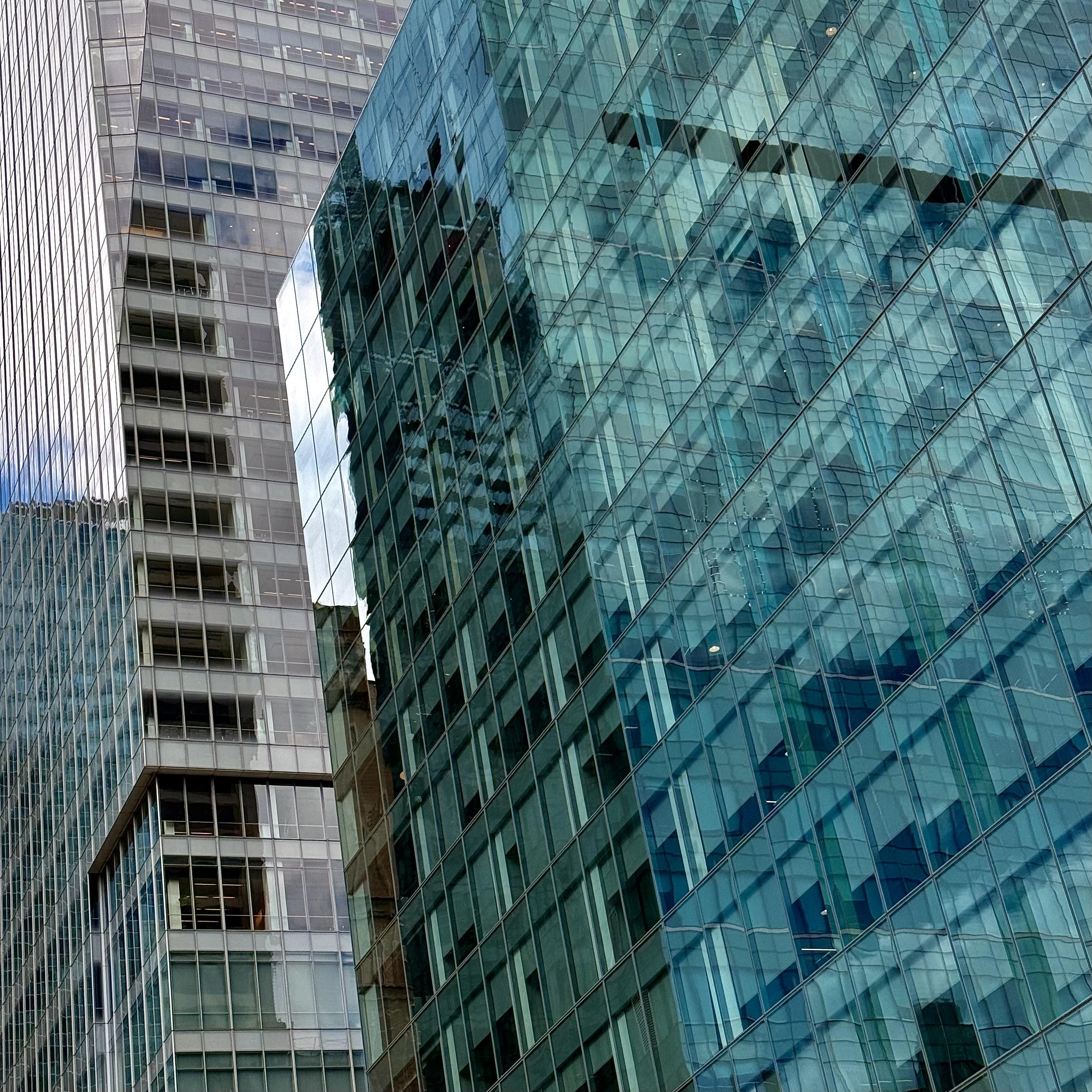 A color, high-angle close-up photograph capturing the textured glass facades of two adjacent skyscrapers. The building on the right features a vibrant, gridded, blue-green glass curtain wall that reflects bright light and distorted shapes of other structures. To its left, a taller, lighter-colored skyscraper with a gray-and-silver glass facade rises, creating a strong contrast in color, texture, and pattern. The lines and reflections create a dynamic, abstract urban composition
