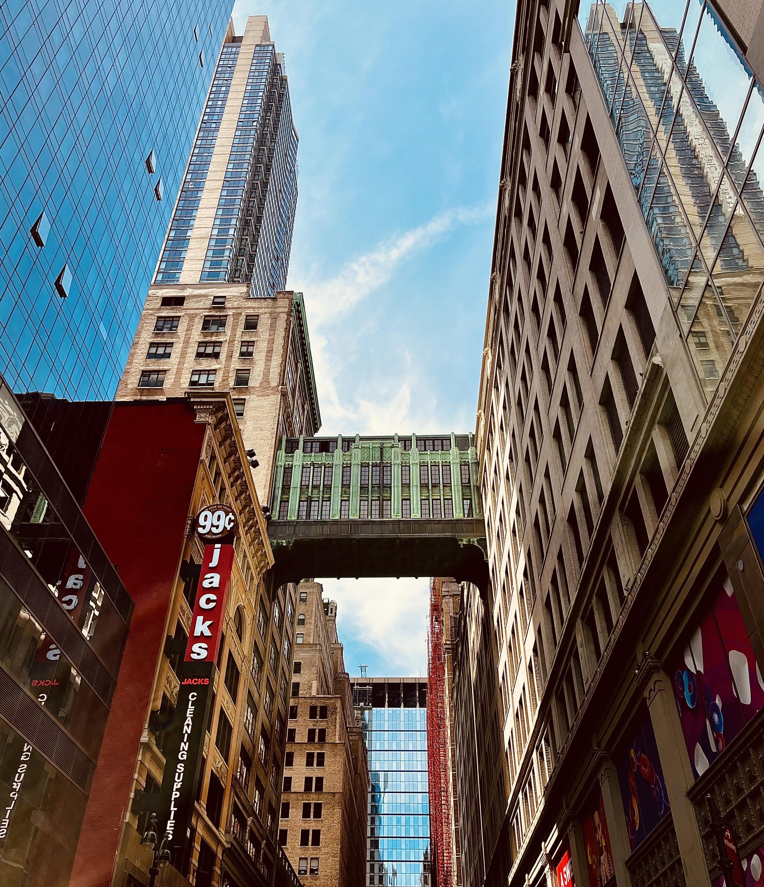 A color, low-angle photograph capturing a narrow street surrounded by skyscrapers under a bright, partially cloudy sky. The view is dominated by a distinct bronze and green skybridge connecting the buildings on either side of the street. A brick building with large vertical signage reading "99¢ Jacks Cleaning Supplies" is partially visible. The modern glass façade of a very tall building is reflected in the glass windows on the right side of the frame. A glass building defines the horizon beneath the skybridge. 