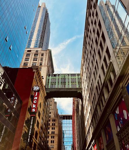 A color, low-angle photograph capturing a narrow street surrounded by skyscrapers under a bright, partially cloudy sky. The view is dominated by a distinct bronze and green skybridge connecting the buildings on either side of the street. A brick building with large vertical signage reading "99¢ Jacks Cleaning Supplies" is partially visible. The modern glass façade of a very tall building is reflected in the glass windows on the right side of the frame. A glass building defines the horizon beneath the skybridge. 