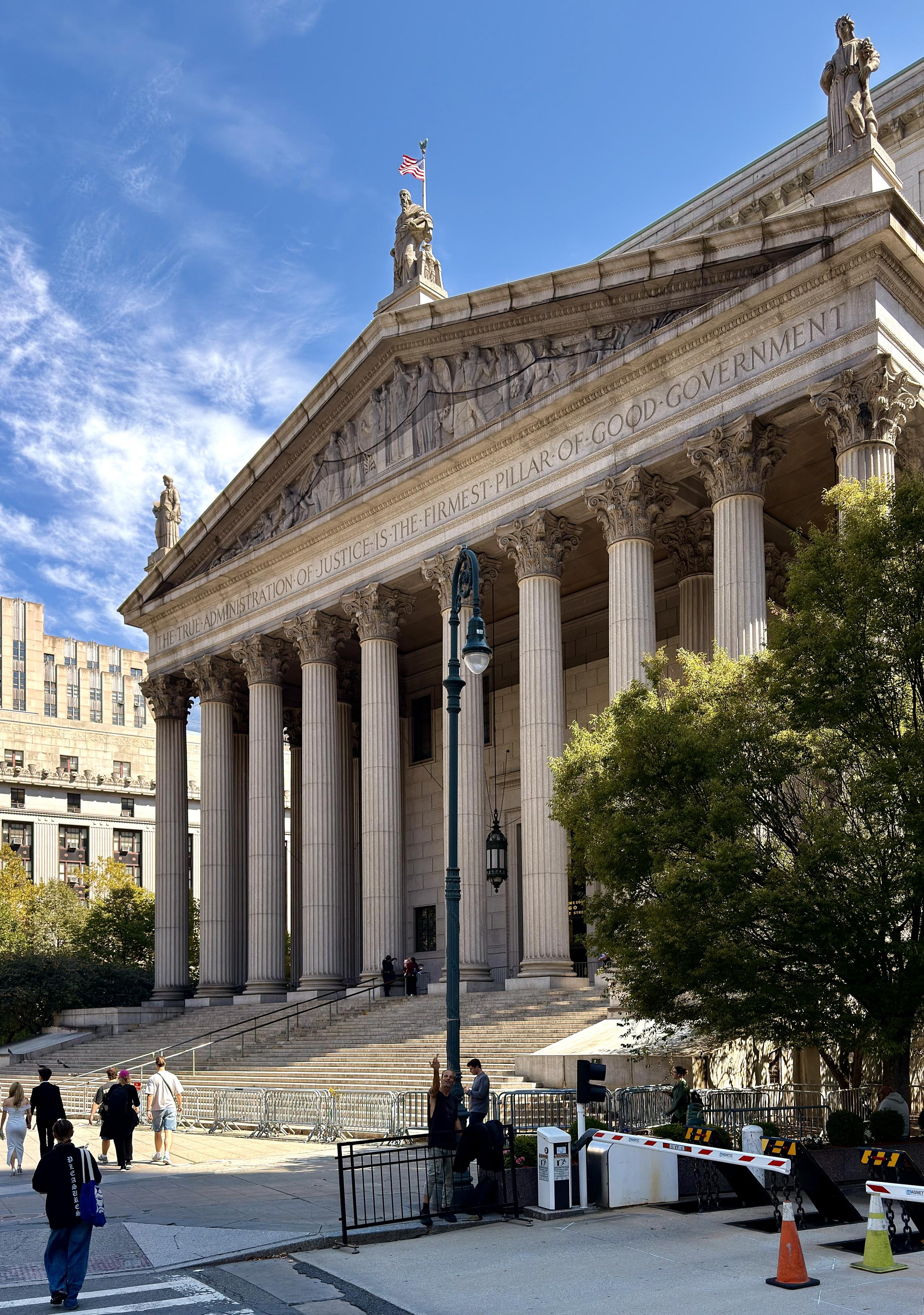 A color photograph taken along Centre Street in the Financial District neighborhood of lower Manhattan. The focal point of the image is the Supreme Court of the State of New York, a monumental neoclassical building with a large triangular pediment supported by a row of tall Corinthian columns. The building's facade is inscribed with the motto, "THE TRUE ADMINISTRATION OF JUSTICE IS THE FIRMEST PILLAR OF GOOD GOVERNMENT". A grand staircase leads up to the main entrance. Sculptures adorn the roofline and pediment. In the foreground, there is a street lamp, a black metal fence with a security barrier, and a large, leafy green tree on the right partially obscuring the building. Several people are visible walking on the steps and the pavement in the lower left and center. A man is standing under the lamp post wearing distressed jeans and a sleeveless black t-shirt. He is facing the camera with his arm in the air giving me the middle finger as I take the shot. The sky above is bright blue with wispy white clouds.