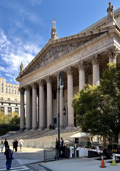 A color photograph taken along Centre Street in the Financial District neighborhood of lower Manhattan. The focal point of the image is the Supreme Court of the State of New York, a monumental neoclassical building with a large triangular pediment supported by a row of tall Corinthian columns. The building's facade is inscribed with the motto, "THE TRUE ADMINISTRATION OF JUSTICE IS THE FIRMEST PILLAR OF GOOD GOVERNMENT". A grand staircase leads up to the main entrance. Sculptures adorn the roofline and pediment. In the foreground, there is a street lamp, a black metal fence with a security barrier, and a large, leafy green tree on the right partially obscuring the building. Several people are visible walking on the steps and the pavement in the lower left and center. A man is standing under the lamp post wearing distressed jeans and a sleeveless black t-shirt. He is facing the camera with his arm in the air giving me the middle finger as I take the shot. The sky above is bright blue with wispy white clouds.