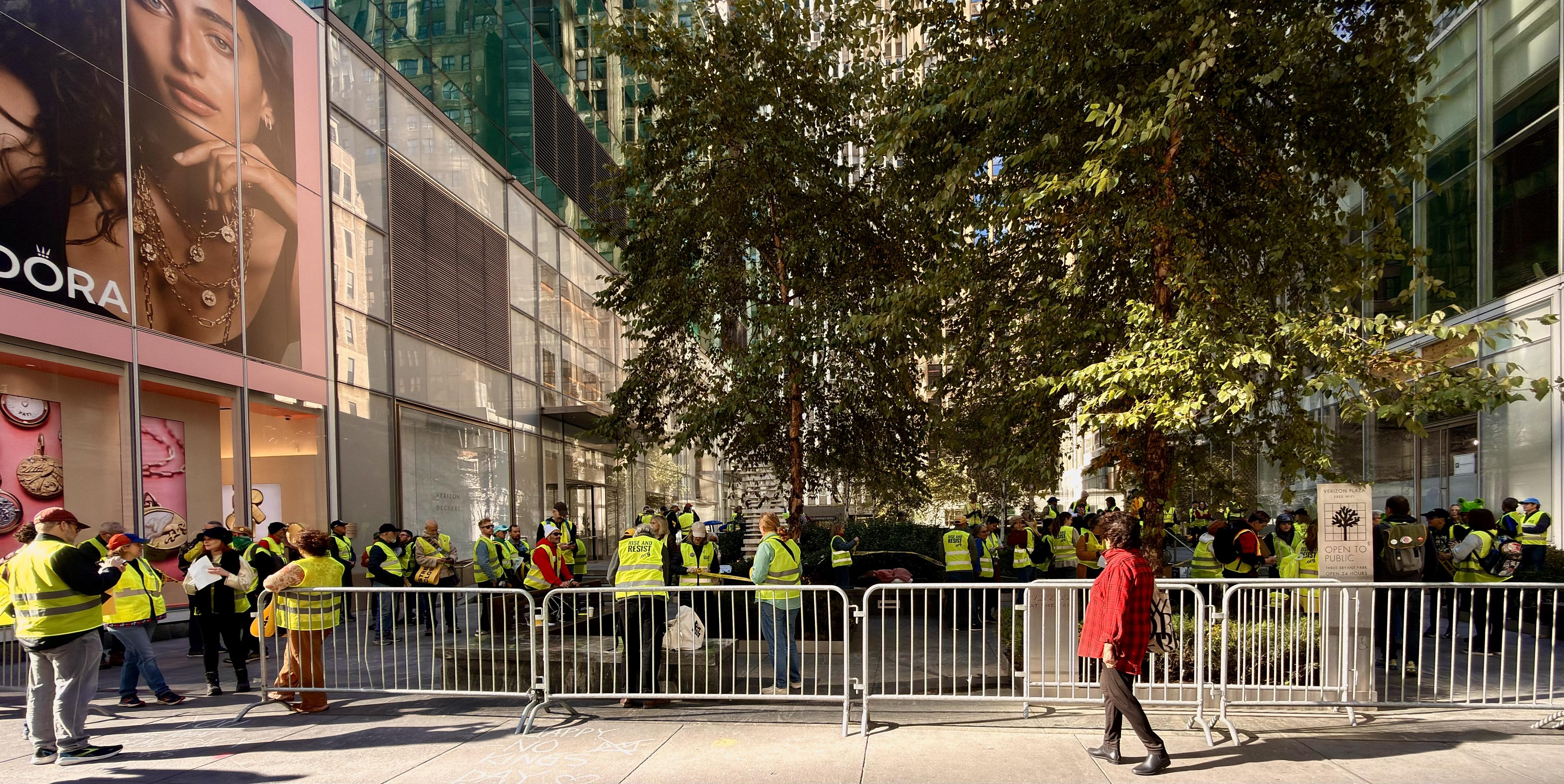A color photograph captures the staging area for No Kings protest volunteers inside Verizon Plaza on a sunny morning. A crowd of people, most of whom are wearing bright yellow-green safety vests, are gathered within a temporary barrier made of interlocking metal barricades. On the left, a large, vertical billboard for "Pandora" jewelry is prominent, featuring a close-up image of a model wearing jewelry. The storefront below has large glass windows. The staging area is defined by the metal barricades running horizontally across the middle ground. Inside the barricades, many people in the yellow-green vests are standing, talking, and looking around. Some are holding clipboards and safety signs. The vests have text on the back "VOLUNTEER" or “RISE AND RESIST. In the center and right background, two large, mature trees with green and yellowing leaves stand within the plaza.