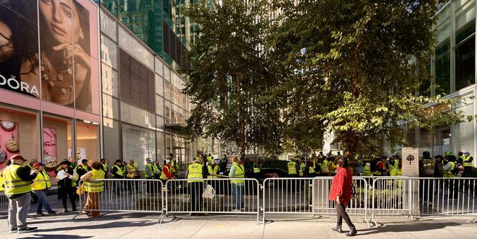 A color photograph captures the staging area for No Kings protest volunteers inside Verizon Plaza on a sunny morning. A crowd of people, most of whom are wearing bright yellow-green safety vests, are gathered within a temporary barrier made of interlocking metal barricades. On the left, a large, vertical billboard for "Pandora" jewelry is prominent, featuring a close-up image of a model wearing jewelry. The storefront below has large glass windows. The staging area is defined by the metal barricades running horizontally across the middle ground. Inside the barricades, many people in the yellow-green vests are standing, talking, and looking around. Some are holding clipboards and safety signs. The vests have text on the back "VOLUNTEER" or “RISE AND RESIST. In the center and right background, two large, mature trees with green and yellowing leaves stand within the plaza.