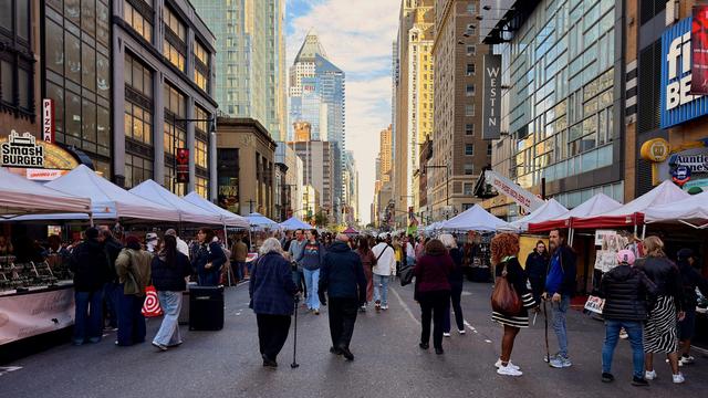 A color, wide angle, eye-level photograph captures a busy street fair on Eighth Avenue in Manhattan. The street is closed to vehicle traffic due to the No Kings protest and is filled with a diverse crowd of people strolling and browsing among rows of white tented vendor stalls lining both sides of the asphalt street. The fair occupies the foreground and middle ground, with people dressed in warm clothing on a crisp morning. Prominent storefronts like Smashburger and a Westin hotel are partially visible behind the temporary tents. The rich blue sky is partly cloudy.