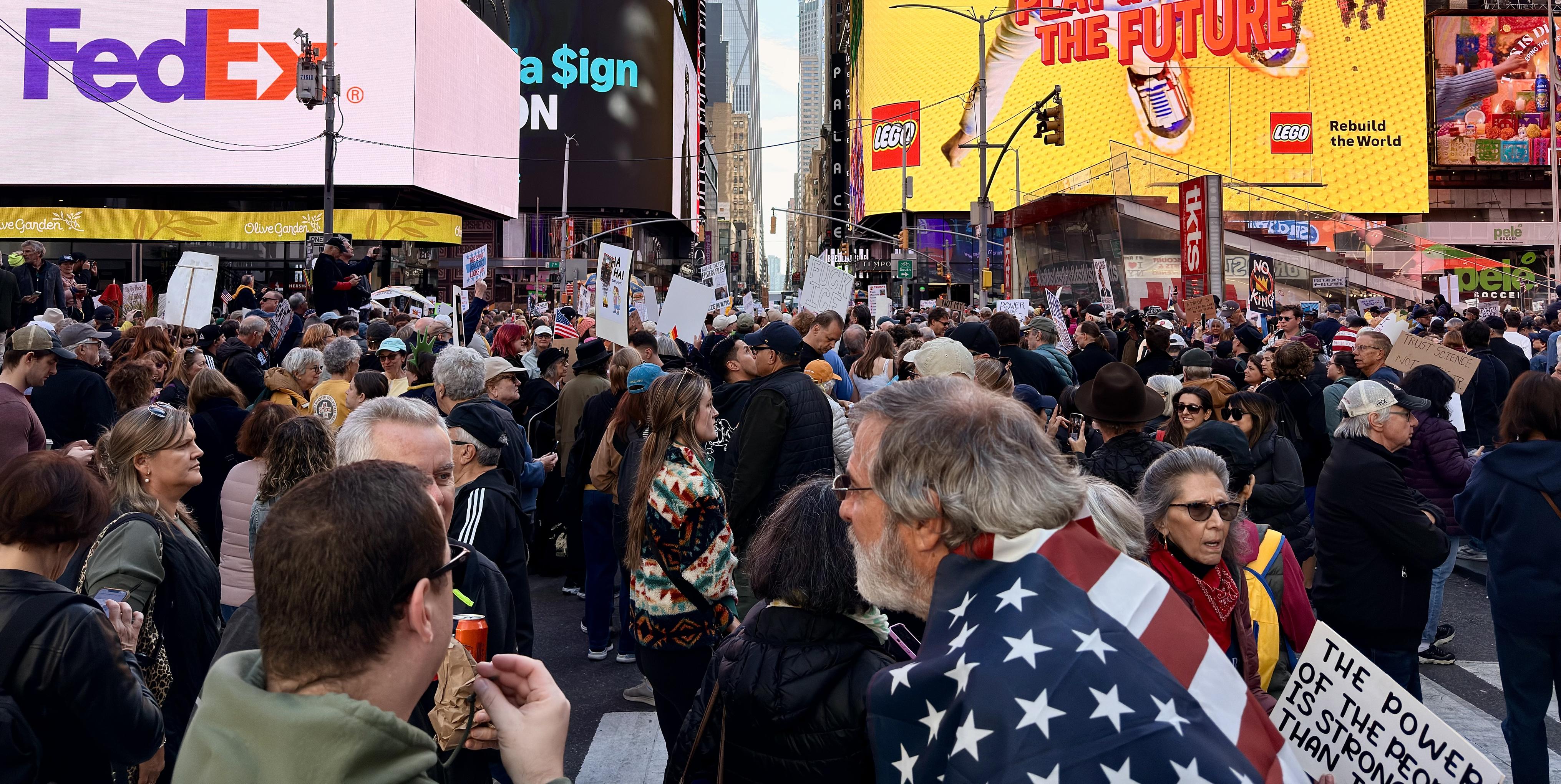 A color, wide angle photograph of a densely packed, diverse crowd of protesters filling the foreground and mid-ground of Father Duffy Square in Times Square, Manhattan. The group consists of people spanning various ages and ethnicities, with many wearing cold-weather clothing like jackets and hats. A man in the immediate foreground, facing slightly away, is draped with an American flag over his shoulders and is looking to the right. The protest takes place amidst the signature brightly lit, massive digital billboards of Times Square. Prominent signage includes a large pink-and-white FedEx logo on a screen to the upper left and, to the upper right, a bright yellow sign with the text "BUILD THE FUTURE" featuring a Lego advertisement with a white sneaker. Traffic lights and typical street infrastructure are visible above the crowd.