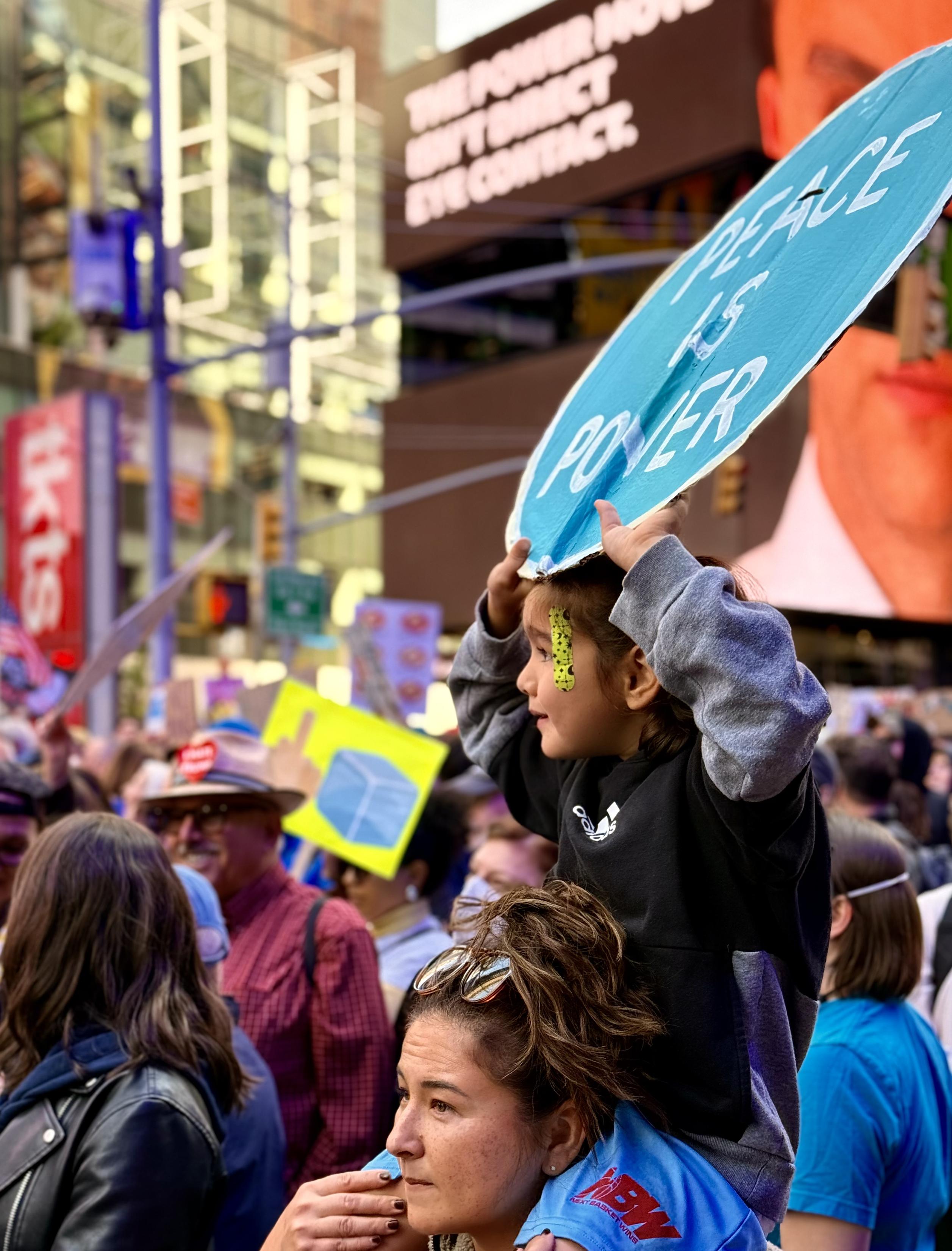 A color photograph in portrait orientation captures a moment at a bustling protest in Times Square, New York City. The focus is on a young child who is sitting on her mother’s shoulders and holding up a homemade, bright blue protest sign. The sign is roughly painted with white capital letters that read, "PEACE IS POWER." The child, wearing a black sweatshirt with a white logo, and blue sport shorts, looks off to the right with a focused expression. A small, yellow-green bandage is on the left side of her face above the cheek. The adult beneath the child is seen from the chest up, with sunglasses resting on her head. The background is a vibrant, dense crowd of demonstrators with signs and hats. Towering above the crowd are the iconic billboards of Times Square.