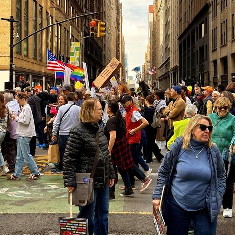A color, street-level photograph of the No Kings protest marching down 7th Avenue in Manhattan. The street is closed to traffic, and the protesters occupy the asphalt, moving between tall buildings that line both sides of the street. The crowd extends into the middle ground and background, demonstrating the scale of the protest. Above the sea of heads, several signs and flags are visible, adding context to the demonstration. Notably, an American flag and a rainbow pride flag are prominently displayed among the signs.