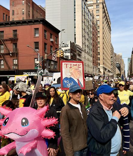 A color photograph captures the No Kings protest marching down Seventh Avenue in Manhattan. The foreground features several distinct individuals amidst a dense crowd of protesters. On the left, a large, bright-pink inflatable axolotl, a type of salamander, with prominent gills and large black eyes. The background provides context for the protest and the location. Protesters are holding various signs, one of which is clearly visible above the center of the crowd, a large red-bordered sign with a portrait of Donald Trump, and the text "TIME to go!". It mimics Time magazine cover. Other signs with the phrase "NO KINGS". The protest is flanked by tall Manhattan buildings; on the left, a low-rise, older brick building with fire escapes is visible above a Taco Bell sign, contrasting with the taller, more modern commercial buildings that line the right side of the avenue under a clear, bright sky.