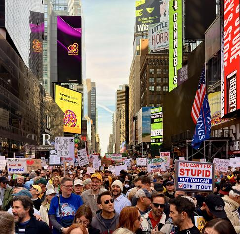 A color, wide-angle, slightly elevated above street level photograph captures a large crowd of people participating in a No Kings protest filling Seventh Avenue in Times Square, Manhattan. The street is packed with demonstrators holding various signs with political messages. The signs are varied, with some prominent ones visible in the foreground, including "NO ONE IS ABOVE THE LAW," "NO HUMAN IS ILLEGAL," "TRUST IS NOT APPEASEMENT," "YOU CAN'T FIX STUPID BUT YOU CAN VOTE IT OUT," and "NO KINGS" on a large blue flag. The crowd is diverse in age and dress, facing generally toward the camera. The background is dominated by the tall, densely packed buildings and bright, illuminated billboards of Times Square. Notable signage includes large advertisements for "That Smooth Silk Taste," "CANNABIS," "HERSHEY'S.” The upper portion of the buildings shows the mix of older stone architecture and modern glass facades. The sky above the buildings is mostly clear with a hazy, bright afternoon light.