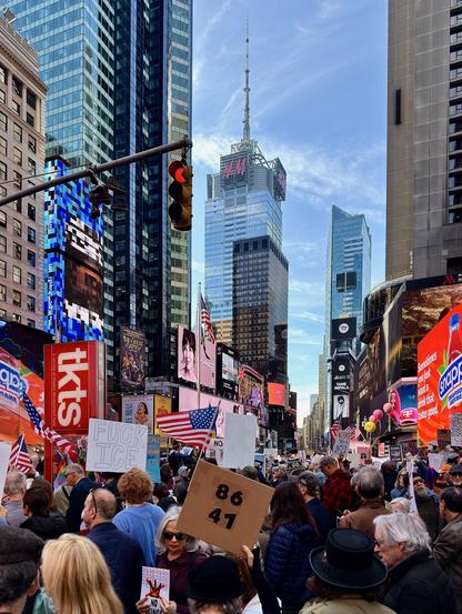 A color, street-level photograph captures a segment of the No Kings protest filling Times Square, Manhattan. The mass of people, largely comprised of adults, extends from the foreground back into the distance, entirely obscuring the street pavement and sidewalks. The crowd is very densely packed and peaceful. Several signs are held by individuals in the foreground. One prominent, square cardboard sign held at the center features large, handwritten black numbers: "86 41.” Other visible signs include a white sign on the left with black text, "FUCK ICE," and a smaller sign in the lower foreground reading, "NO KINGS." American flags are interspersed throughout the crowd, carried on poles or worn on clothing. The backdrop of the scene is dominated by tall buildings and advertising. Visible structures include the H&M building topped by a spire and the flashing digital signs for "tktS," Snapple, and various entertainment billboards in the middle and right background. The sky above is a bright blue with some wispy clouds. A traffic light on the left is illuminated with a red signal.