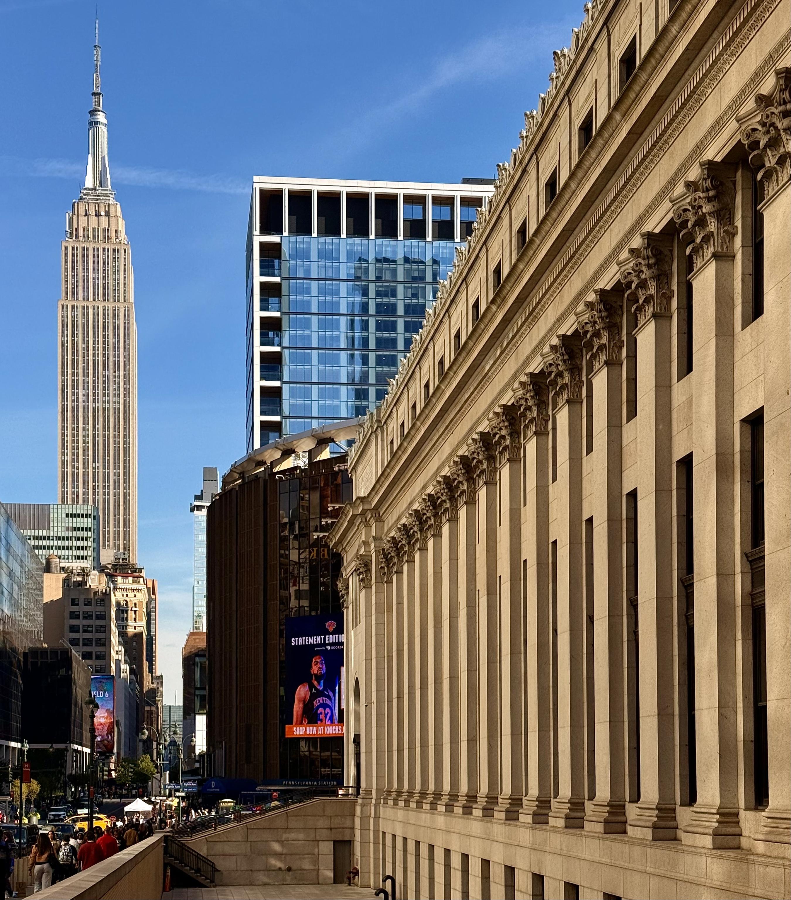 A color photograph captures an architectural tableau in the Chelsea neighborhood of Manhattan on a bright, sunny day with a clear blue sky. On the far left, the Empire State Building, its classic Art Deco spire a stark vertical presence. The foreground is anchored on the right by Moynihan Train Hall. Its long row of colossal columns and intricate classical details provide a strong horizontal and historical counterpoint. Between these two landmarks, a modern, glass and white-framed high-rise stands, reflecting the blue sky. The middle ground also features a large, dark, cylindrical structure, which is part of the Madison Square Garden complex. A large digital banner on this building displays an advertisement. The bottom left shows a busy street scene with people and traffic.