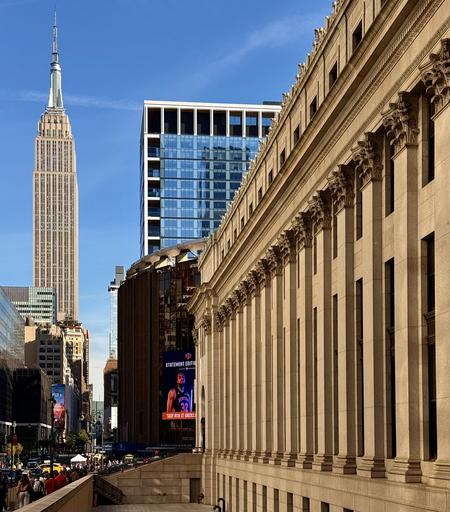 A color photograph captures an architectural tableau in the Chelsea neighborhood of Manhattan on a bright, sunny day with a clear blue sky. On the far left, the Empire State Building, its classic Art Deco spire a stark vertical presence. The foreground is anchored on the right by Moynihan Train Hall. Its long row of colossal columns and intricate classical details provide a strong horizontal and historical counterpoint. Between these two landmarks, a modern, glass and white-framed high-rise stands, reflecting the blue sky. The middle ground also features a large, dark, cylindrical structure, which is part of the Madison Square Garden complex. A large digital banner on this building displays an advertisement. The bottom left shows a busy street scene with people and traffic.