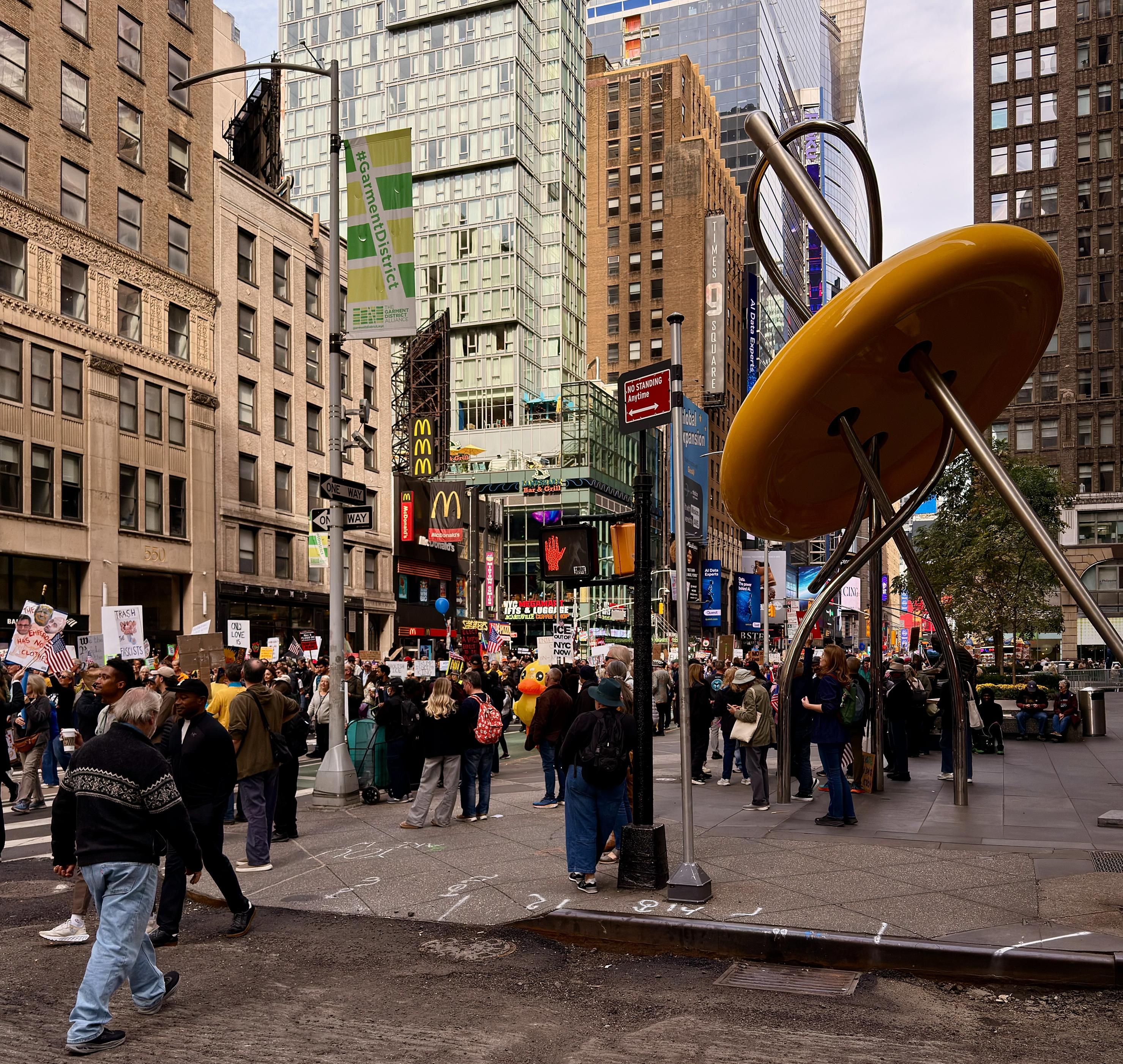 A color photograph captures the No Kings protest marching down Seventh Avenue in the Garment District neighborhood of Manhattan. A thick crowd of demonstrators, many holding signs and flags, fills the left side and center of the frame as they move down the street. The urban environment is marked by tall office and residential buildings, including one with a "Garment District" banner hanging from its facade. In the foreground, dominating the right side, is a striking piece of public art: a colossal, bright yellow sculpture resembling a button on a pedestal, which is the "Needle Threading A Button" art installation.