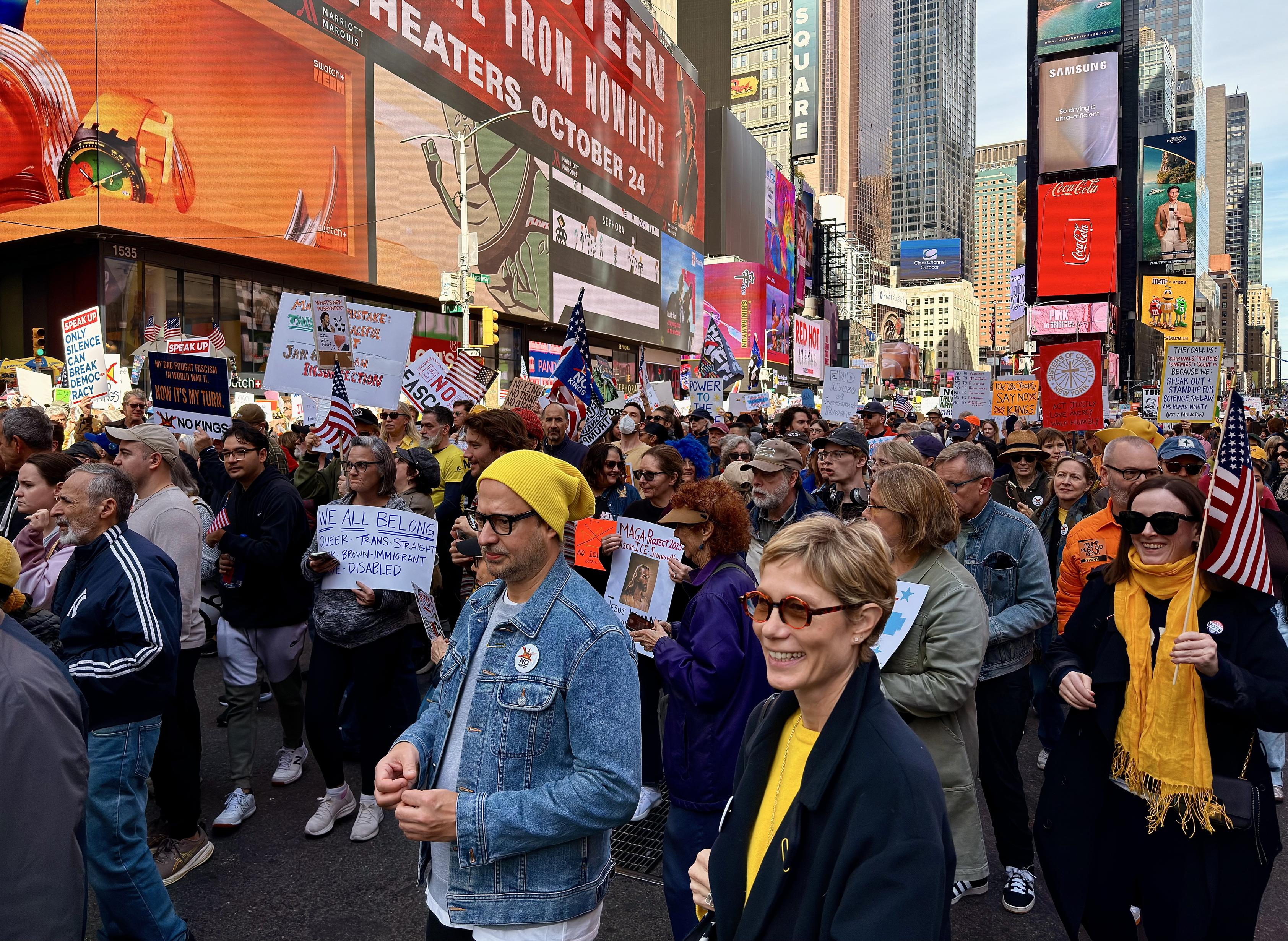 A color, street-level photograph capturing a wide view of a crowded political demonstration in Times Square, Manhattan for the No Kings protest. A diverse group of people fill the streets and sidewalk, many holding signs and American flags. The signs visible carry various messages, including “FUCK ICE,” “FUCK TRUMP,” and "NO KINGS." In the foreground, a woman with short blonde hair and sunglasses is smiling, wearing a dark blue coat over a bright yellow top, with a man in a denim jacket and a yellow beanie next to her. The background is dominated by the tall buildings and bright, large digital billboards characteristic of Times Square. The sky above is clear and bright. The crowd is facing the camera’s direction, moving slowly and peacefully south.