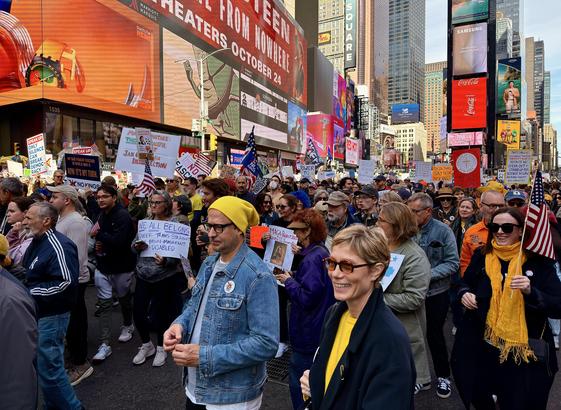 A color, street-level photograph capturing a wide view of a crowded political demonstration in Times Square, Manhattan for the No Kings protest. A diverse group of people fill the streets and sidewalk, many holding signs and American flags. The signs visible carry various messages, including “FUCK ICE,” “FUCK TRUMP,” and "NO KINGS." In the foreground, a woman with short blonde hair and sunglasses is smiling, wearing a dark blue coat over a bright yellow top, with a man in a denim jacket and a yellow beanie next to her. The background is dominated by the tall buildings and bright, large digital billboards characteristic of Times Square. The sky above is clear and bright. The crowd is facing the camera’s direction, moving slowly and peacefully south.