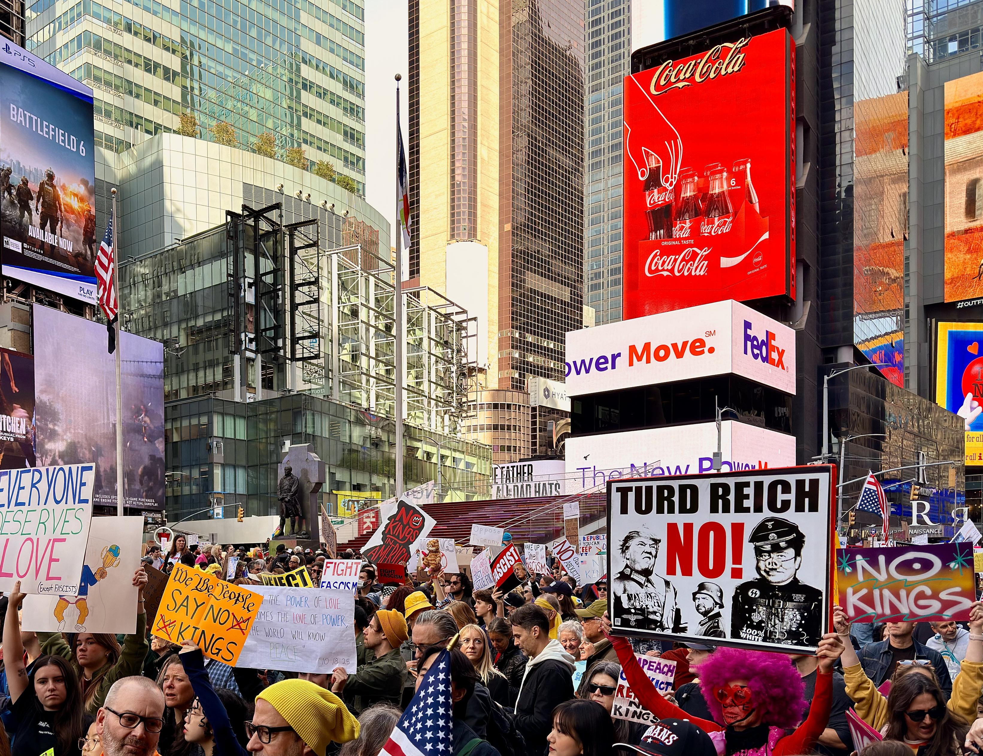 A color, medium-angle, street-level photograph captures the massive No Kings protest filling the main pedestrian area of Times Square in New York City, set against a backdrop of towering, modern skyscrapers adorned with giant billboards. Thousands of diverse protestors are tightly packed together, holding various hand-made signs, with the crowd extending deep into the background. Dominating the lower-right foreground is a large, square protest sign with the bold, all-caps text: "TURD REICH NO!" And Donald Trump, JD Vance, and Elon Musk are dressed as SS nazi soldiers below the text. Other visible signs include a yellow one reading: "We The People Say NO KINGS," another saying: "EVERYONE DESERVES LOVE," and a red sign on the far right that reiterates "NO KINGS". Above the crowd, the iconic, brightly lit billboards are visible, including a dominant red Coca-Cola sign on a building to the right, and a large 'BATTLEFIELD 6' PlayStation advertisement banner on the left.