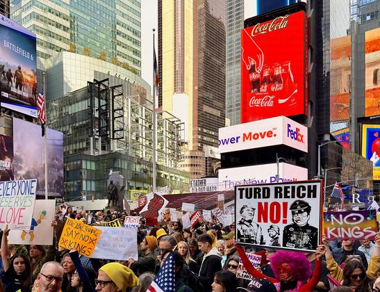 A color, medium-angle, street-level photograph captures the massive No Kings protest filling the main pedestrian area of Times Square in New York City, set against a backdrop of towering, modern skyscrapers adorned with giant billboards. Thousands of diverse protestors are tightly packed together, holding various hand-made signs, with the crowd extending deep into the background. Dominating the lower-right foreground is a large, square protest sign with the bold, all-caps text: "TURD REICH NO!" And Donald Trump, JD Vance, and Elon Musk are dressed as SS nazi soldiers below the text. Other visible signs include a yellow one reading: "We The People Say NO KINGS," another saying: "EVERYONE DESERVES LOVE," and a red sign on the far right that reiterates "NO KINGS". Above the crowd, the iconic, brightly lit billboards are visible, including a dominant red Coca-Cola sign on a building to the right, and a large 'BATTLEFIELD 6' PlayStation advertisement banner on the left.