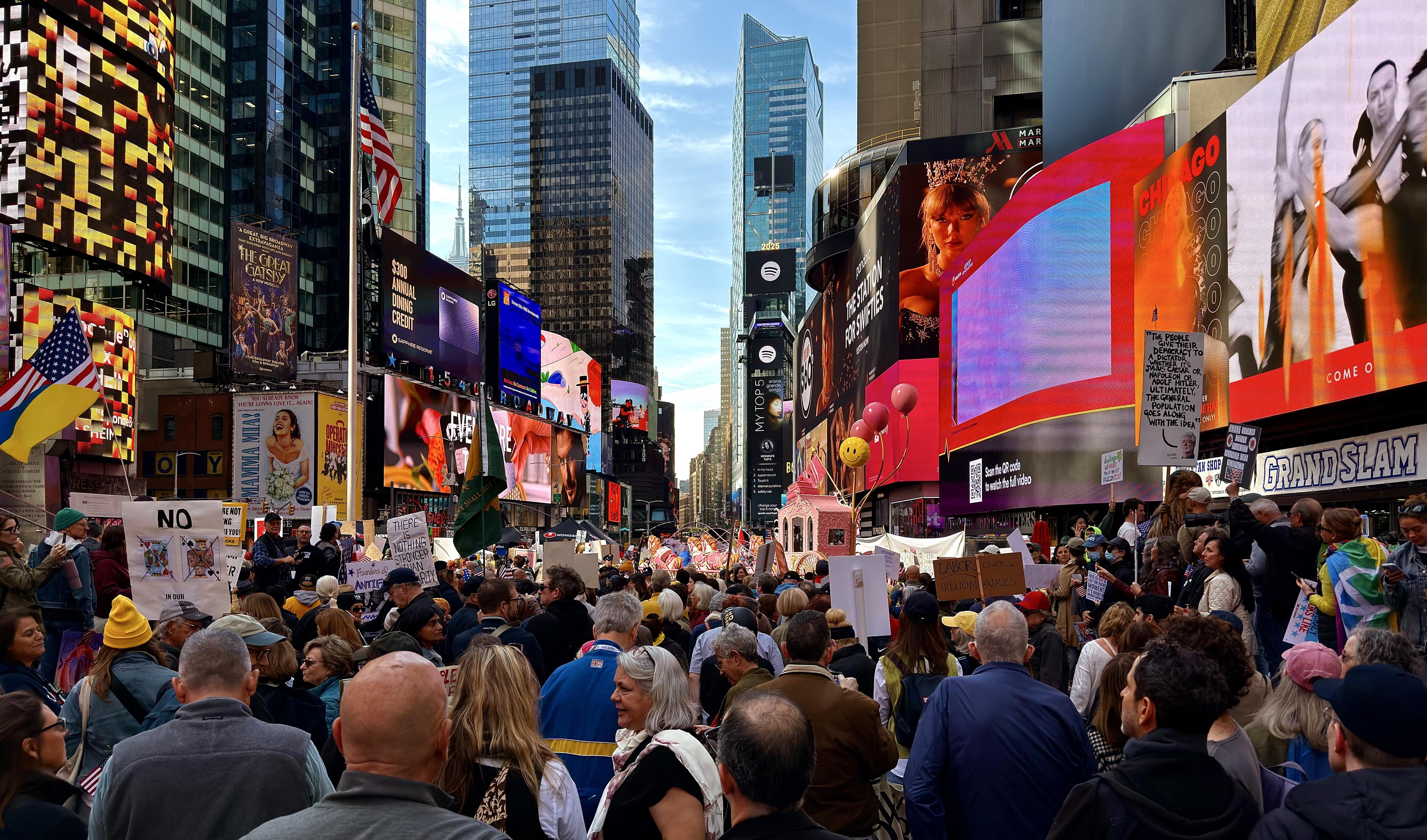 A color, wide-angle, street-level photograph of the No Kings protest in Times Square as protestors begin to start marching down Seventh Avenue. The perspective is from behind the frontmost part of the crowd. The protesters are a diverse mix of people, primarily focused away from the camera. They hold various signs and flags, though many are obscured by the sheer number of people. The backdrop is commercial advertising, characteristic of Times Square. Towering above the protesters on the right is a massive digital billboard featuring a close-up image of Taylor Swift's face. Other screens display brightly colored abstract patterns, commercials, and public service messages. The street itself is completely taken over by the gathering. The sky above is clear and light blue.