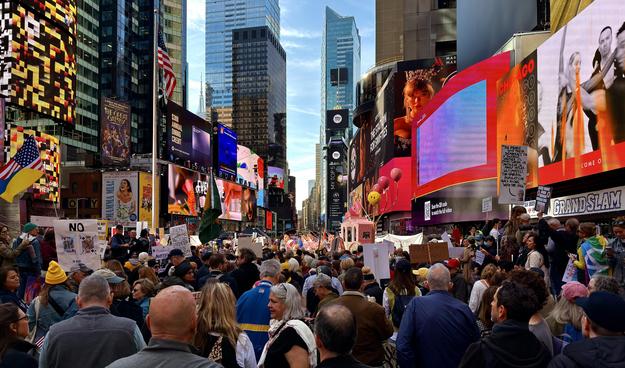 A color, wide-angle, street-level photograph of the No Kings protest in Times Square as protestors begin to start marching down Seventh Avenue. The perspective is from behind the frontmost part of the crowd. The protesters are a diverse mix of people, primarily focused away from the camera. They hold various signs and flags, though many are obscured by the sheer number of people. The backdrop is commercial advertising, characteristic of Times Square. Towering above the protesters on the right is a massive digital billboard featuring a close-up image of Taylor Swift's face. Other screens display brightly colored abstract patterns, commercials, and public service messages. The street itself is completely taken over by the gathering. The sky above is clear and light blue.