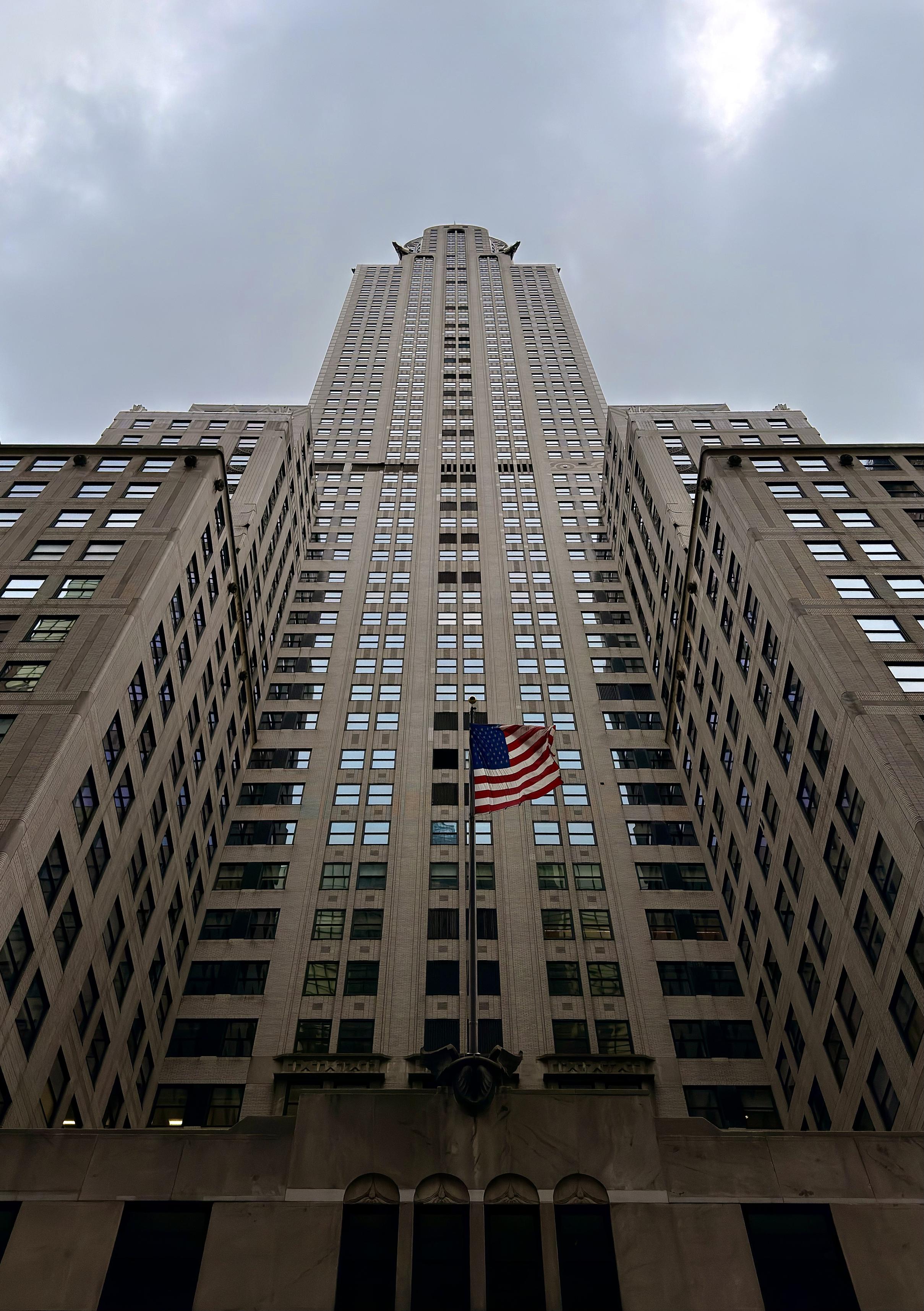 A color, low-angle, portrait orientation photograph if the full verticality of the Chrysler Building in midtown Manhattan on an overcast day. The view is centered directly on the building's main shaft, emphasizing its tremendous height and the continuous vertical lines of its facade. The building's Art Deco style is evident in the geometric repetition of the numerous small windows and the stepped, monumental presence of the lower stories that frame the central tower. The stone-colored exterior and the subdued light of the cloudy sky give the image a slightly desaturated, imposing atmosphere. Prominently featured near the base of the central shaft is a large American flag mounted on a flagpole, fluttering gently in front of the building's entrance. The red, white, and blue of the flag provides a striking burst of color against the neutral tones of the architecture and the gray sky. The perspective, taken from very close to the ground, makes the skyscraper appear to soar endlessly into the cloudy sky, creating a powerful sense of scale and architectural grandeur.