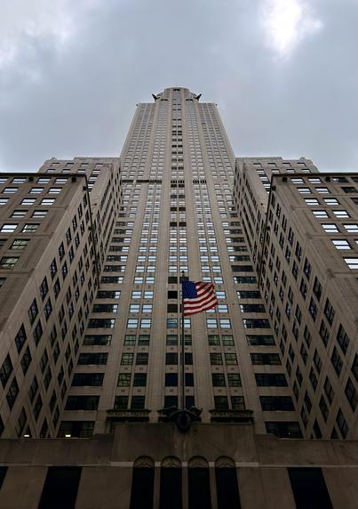 A color, low-angle, portrait orientation photograph if the full verticality of the Chrysler Building in midtown Manhattan on an overcast day. The view is centered directly on the building's main shaft, emphasizing its tremendous height and the continuous vertical lines of its facade. The building's Art Deco style is evident in the geometric repetition of the numerous small windows and the stepped, monumental presence of the lower stories that frame the central tower. The stone-colored exterior and the subdued light of the cloudy sky give the image a slightly desaturated, imposing atmosphere. Prominently featured near the base of the central shaft is a large American flag mounted on a flagpole, fluttering gently in front of the building's entrance. The red, white, and blue of the flag provides a striking burst of color against the neutral tones of the architecture and the gray sky. The perspective, taken from very close to the ground, makes the skyscraper appear to soar endlessly into the cloudy sky, creating a powerful sense of scale and architectural grandeur.