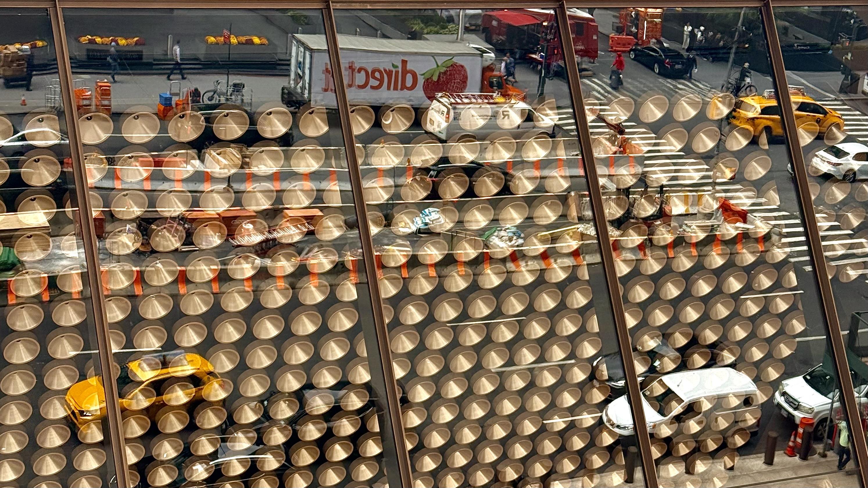 A color, landscape orientation photograph that captures an abstract and busy reflection of Park Avenue on the exterior glass of the JPMorgan Chase building. The street scene is distorted and fragmented by the building's facade, which is covered in a dense, repeating pattern of circular, copper-toned elements. These architectural features create hundreds of overlapping circular highlights and shadows, breaking the reflected image of the street below into countless small, almost lens-like segments. The reflection includes yellow taxi cabs, white cars, a crosswalk, traffic, and pedestrians, all visible through the patterned glass. The reflected street scene contrasts sharply with the static, geometric pattern of the building's exterior. Prominently reflected is a large white truck with red lettering that appears reversed, and red food stands are also visible. The upper portion of the image shows the street level and a brighter, slightly less distorted reflection, while the lower portion is dominated by the repeating circular pattern.