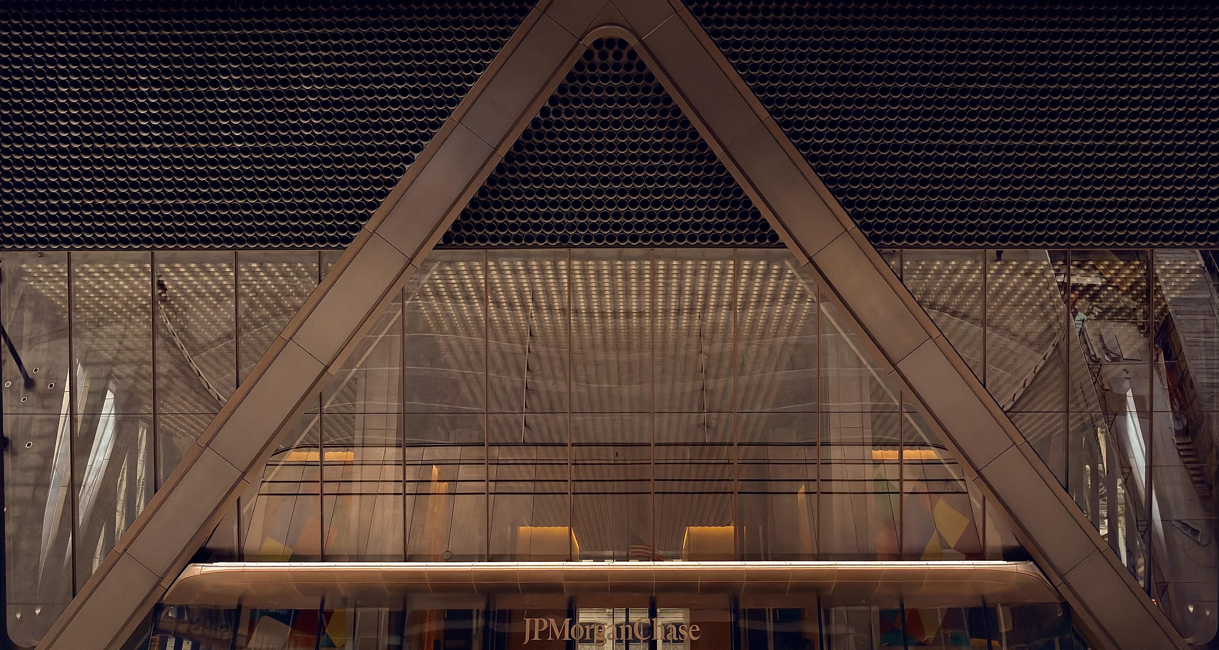 A color photograph on landscape orientation captures the entrance of the JPMorgan & Chase headquarters at 270 Park Avenue. Dominating the center is a massive, inverted 'V' shape formed by thick, light-brown metal beams, which frames the glass-clad lower portion of the skyscraper. This structural 'V' creates a strong, symmetrical, and almost pyramidal composition. Above the 'V' and extending across the top of the frame is a dark, circular, gridded texture that fills the building's facade, creating a densely patterned backdrop. Below the main 'V' structure, the entrance area features multiple layers of clear glass, revealing a spacious, well-lit interior lobby with polished bronze surfaces. The lighting inside is a warm and golden color, and is casting subtle geometric shadows and reflections across the ceiling and walls visible through the glass. Along the very bottom of the image, centered below the main structure, the text "JPMorgan Chase" is visible in a light-colored golden font. The bottom edge also hints at a colorful, abstract pattern artwork visible through the ground-level windows, contrasting with the building's otherwise sleek, dark, and metallic palette.