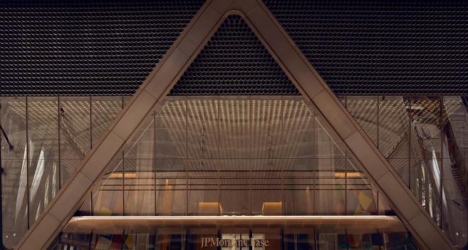 A color photograph on landscape orientation captures the entrance of the JPMorgan & Chase headquarters at 270 Park Avenue. Dominating the center is a massive, inverted 'V' shape formed by thick, light-brown metal beams, which frames the glass-clad lower portion of the skyscraper. This structural 'V' creates a strong, symmetrical, and almost pyramidal composition. Above the 'V' and extending across the top of the frame is a dark, circular, gridded texture that fills the building's facade, creating a densely patterned backdrop. Below the main 'V' structure, the entrance area features multiple layers of clear glass, revealing a spacious, well-lit interior lobby with polished bronze surfaces. The lighting inside is a warm and golden color, and is casting subtle geometric shadows and reflections across the ceiling and walls visible through the glass. Along the very bottom of the image, centered below the main structure, the text "JPMorgan Chase" is visible in a light-colored golden font. The bottom edge also hints at a colorful, abstract pattern artwork visible through the ground-level windows, contrasting with the building's otherwise sleek, dark, and metallic palette.