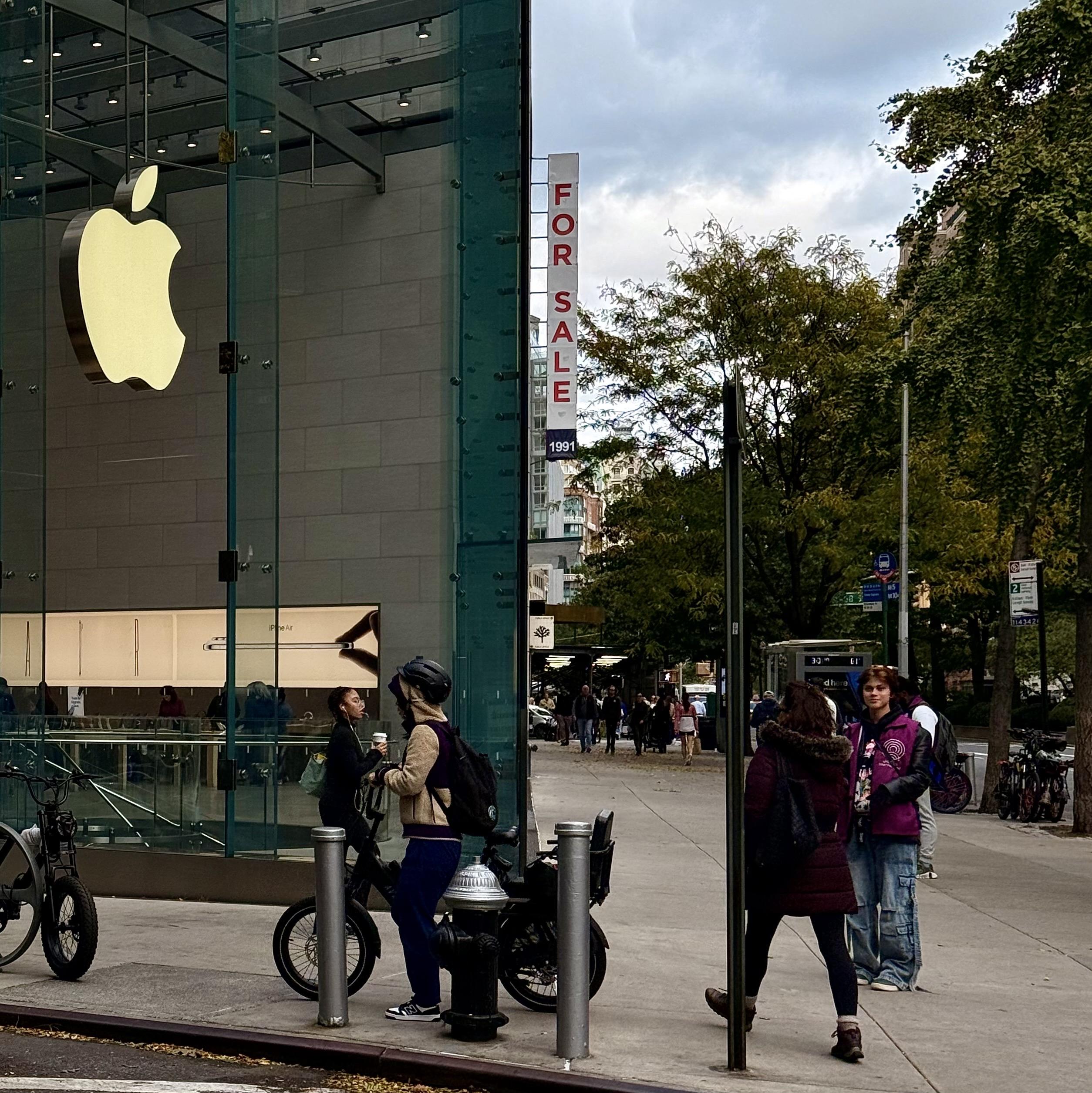 A color photograph in square aspect ratio, taken street-level along Broadway at the intersection with 67th Street in Manhattan. The focal point of the image is the all-glass Apple Store. The address of the store is 1981, and just past the store is 1991 Broadway with a large white vertical sign with red letters that read ‘FOR SALE’. From the vantage point of the camera it looks like the sign is attached to the Apple Store. People are walking and standing on the sidewalk, a person is stopped, standing with a bicycle. Another bicycle is parked in front of them. There are green trees lining the street, and the cloudy afternoon sky fills the frame in the distance.