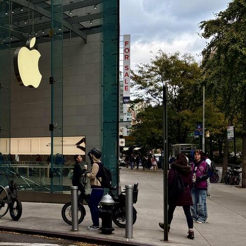 A color photograph in square aspect ratio, taken street-level along Broadway at the intersection with 67th Street in Manhattan. The focal point of the image is the all-glass Apple Store. The address of the store is 1981, and just past the store is 1991 Broadway with a large white vertical sign with red letters that read ‘FOR SALE’. From the vantage point of the camera it looks like the sign is attached to the Apple Store. People are walking and standing on the sidewalk, a person is stopped, standing with a bicycle. Another bicycle is parked in front of them. There are green trees lining the street, and the cloudy afternoon sky fills the frame in the distance.