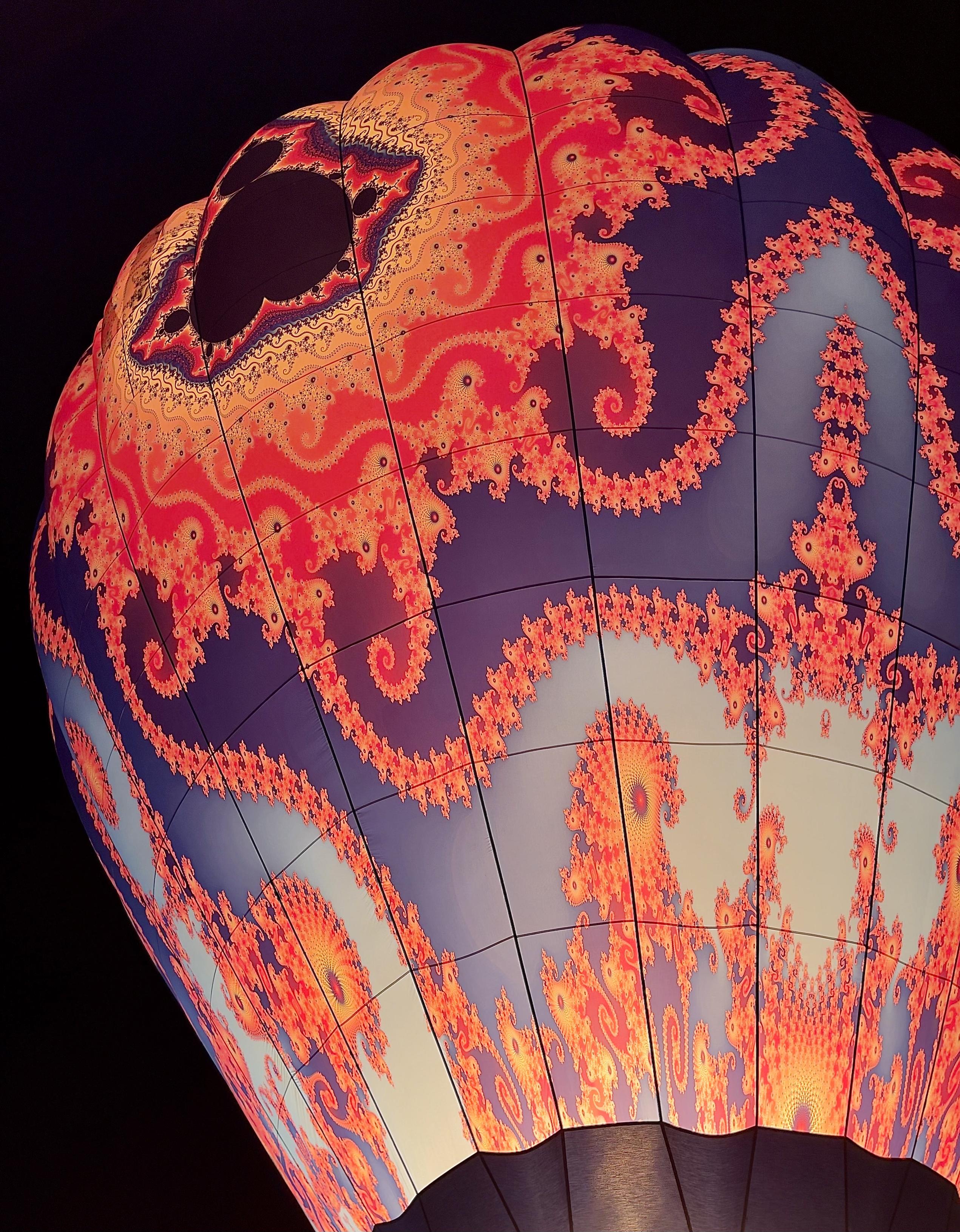 This is a close-up, low-angle photograph of a hot air balloon illuminated from within at night, showing a vibrant and intricate design on its envelope. The pattern is a complex, psychedelic, deep fractal design, primarily in glowing shades of fiery orange, red, and yellow against a dark, cool-toned background of deep indigo and violet. The fractal pattern features countless swirling, self-similar spirals that seem to climb and cascade down the fabric of the balloon. The internal illumination makes the pattern appear to pulse and glow intensely against the dark night sky, drawing attention to the fine, delicate details of the artwork. The perspective captures a significant portion of the balloon's upper hemisphere, emphasizing its massive, curved form and the fabric's texture where the light shines through. The structure is defined by dark, vertical seams running down the balloon. A small portion of the darker material near the base is visible at the very bottom. The contrasting warmth of the fractal pattern and the cool, dark background create a stunning visual effect.