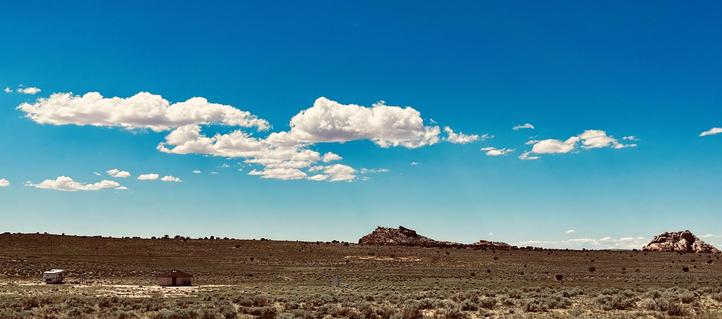 A color panorama photo of the high desert landscape in Navajo Nation, about an hour from Page, Arizona. Light green and brown short desert scrub fills the rolling landscape. In the mid foreground is a small white trailer, a wooden outhouse, and a Navajo hogan dwelling. Further in the distance are two large, pointed rock formations which were used to direct me to the location by the home owner. A few puffy white clouds are passing over the scene in a bright blue sky. 