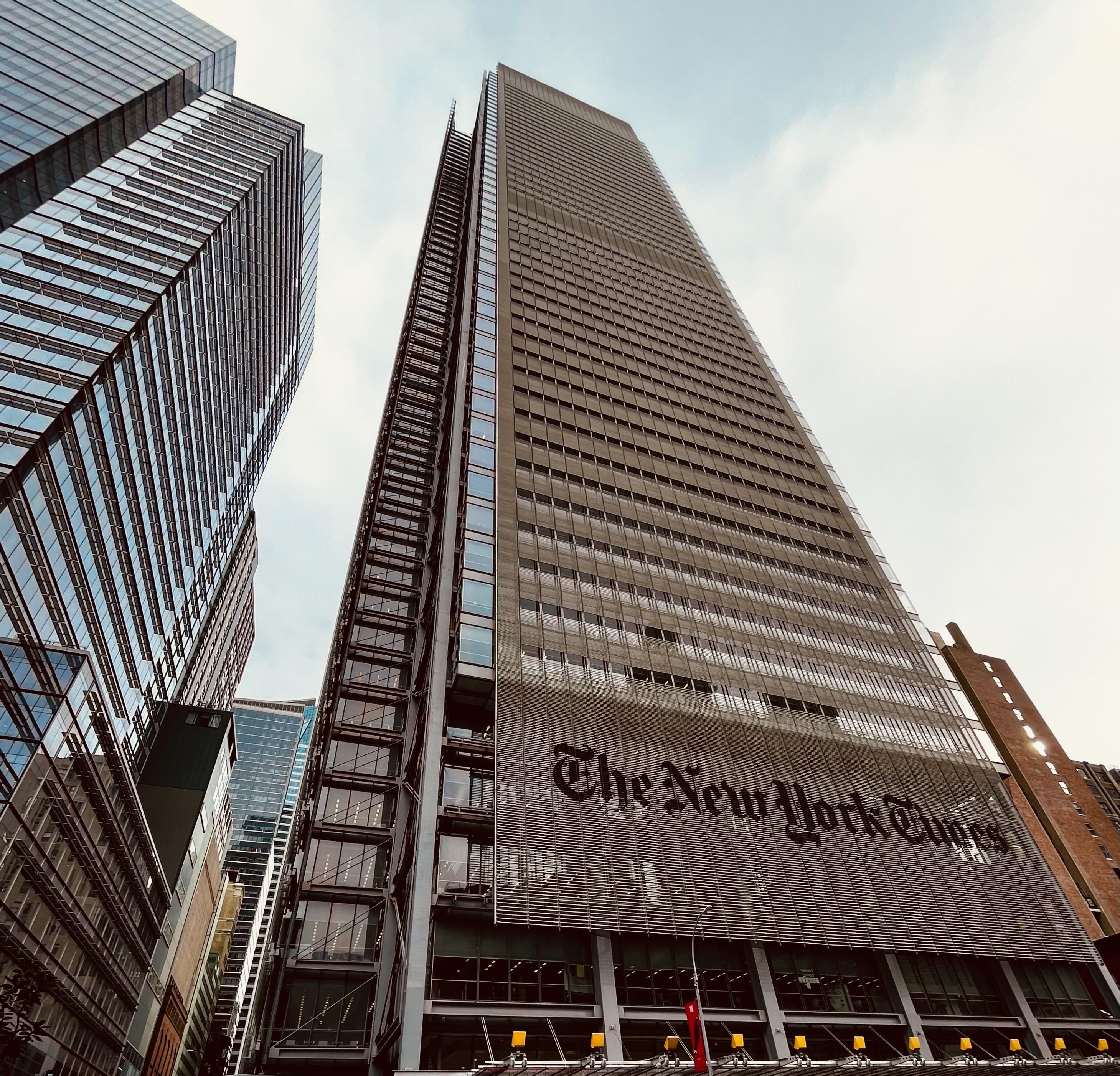 A color photograph in square aspect ratio of the New York Times tower, a tall skyscraper in Manhattan. The photo captures the entire height of the tower as it tapers slightly against a bright, light blue sky with scattered white clouds. The building's facade is notable for its steel-and-glass curtain wall, and a large, stylized "The New York Times" logo is prominently displayed in dark lettering across the lower portion of the visible exterior, set against the light vertical louvers that partially screen the glass. The composition is framed by the adjacent buildings, one of which is a much starker, dark glass structure on the left that reflects the sky and the surrounding city. The bottom of the image shows a high street level, with the roof of the building’s entry visible. The street and sidewalks are not visible.